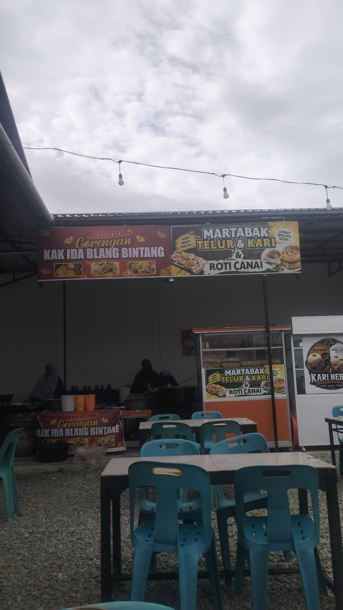 The image depicts an outdoor food stall area, likely in Cotcut, Indonesia, characterized by gravel flooring and covered seating. Overhead, a string of bare light bulbs hangs against a cloudy sky. In the background, a white building with a dark roof provides shelter. Two prominent banners advertise food items. The left banner, in red, reads "TERSEDIA GORENGAN KAK IDA BLANG BINTANG" with images of fried food. The right banner, in black and yellow, advertises "MARTABAK TELUR & KARI" and "ROTI CANAI," also featuring accompanying food imagery.
In the middle ground, a food stall with glass display cases is visible, showcasing eggs and printed advertisements for "MARTABAK TELUR & KARI" and "ROTI CANAI." To the right of this stall is another food stand with a banner for "KARI BEBEK." Two individuals are partially visible behind a counter, appearing to be preparing or serving food. One is wearing a hijab.
The foreground features several tables with light-colored tops and dark legs, surrounded by blue plastic chairs. Some chairs are empty, while others appear to be occupied by the backs of people further within the seating area. The overall scene suggests a casual dining environment at a food court or street food establishment.