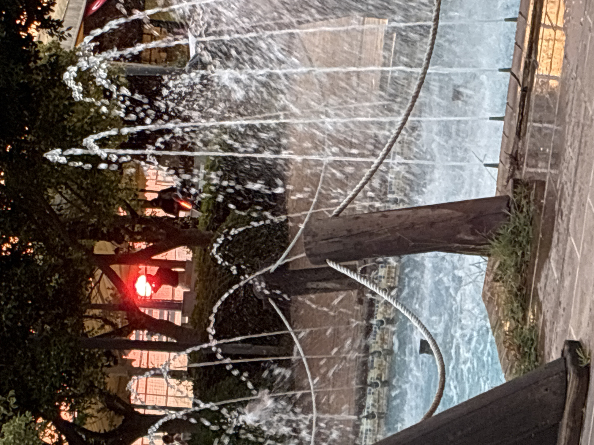 An outdoor fountain is in the foreground, with jets of water spraying upwards and outwards. The water creates a misty effect, obscuring the background. In the middle ground, a thick rope is strung between wooden posts, serving as a barrier. Beyond the rope, the water in the fountain's basin is a vibrant blue. In the background, the setting sun casts a warm, orange glow through the dark silhouettes of trees. A person is visible in the distance, their outline dark against the bright sky, holding what appears to be the sun in their hands, as if cradling it. The scene is likely in a public park or promenade. The image does not contain specific landmarks to pinpoint its location in Alanya, Turkey.