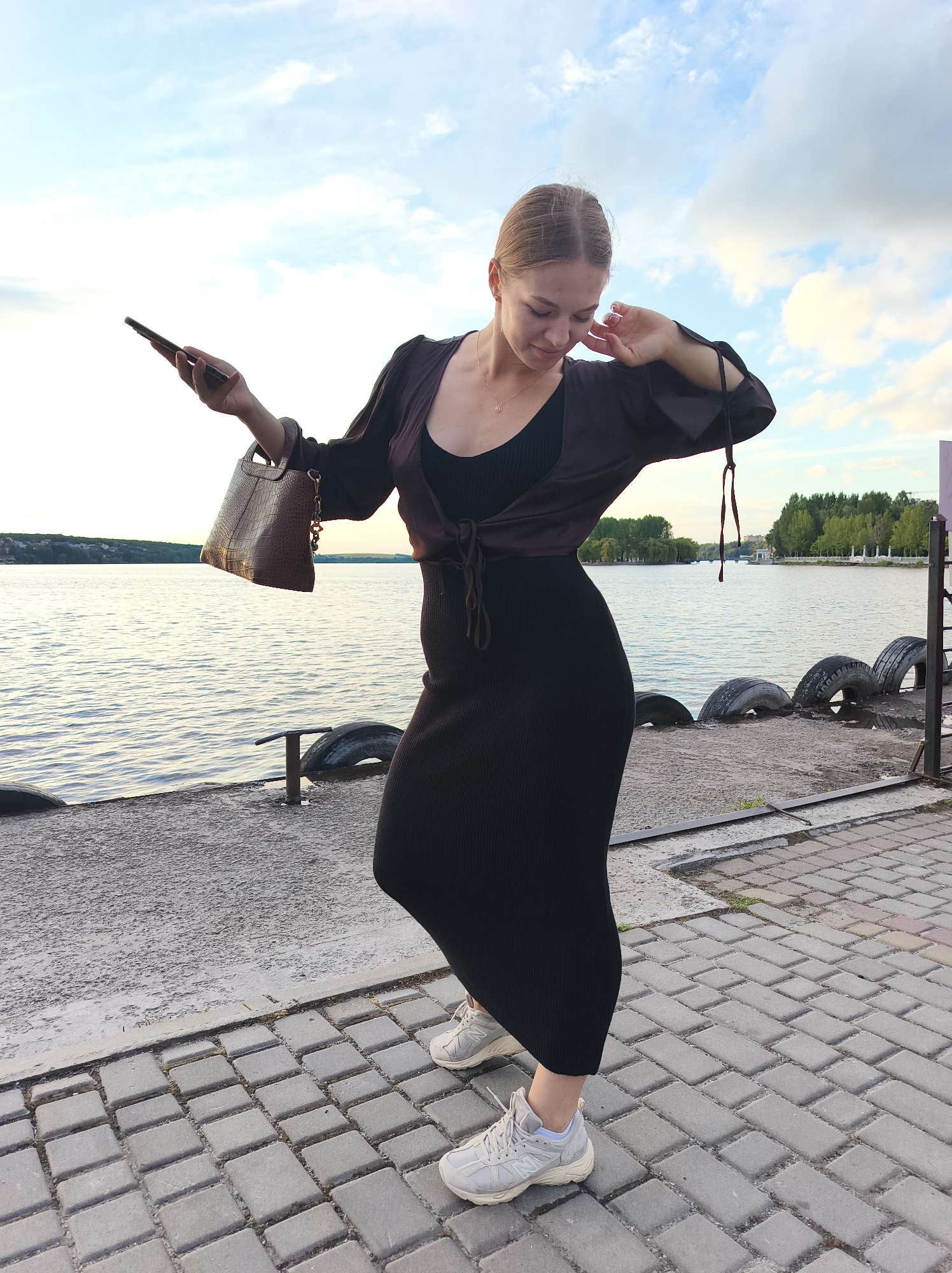A young woman with blonde hair pulled back stands on a paved walkway next to a body of water. She is wearing a long black ribbed dress, a dark brown wrap top tied at the waist, and light-colored athletic shoes. In her right hand, she holds a dark smartphone. Her left hand is raised near her face, and she is looking down. She is also carrying a brown crocodile-embossed handbag. The background shows a calm body of water, a line of trees on the opposite shore, and a partly cloudy sky. Several old tires are visible along the water's edge. The paved walkway is made of light gray rectangular bricks. The scene appears to be a waterfront promenade. This location could potentially be on the Ternopil pond in Ternopil, Ukraine, given the presence of a large pond and urban park-like scenery.