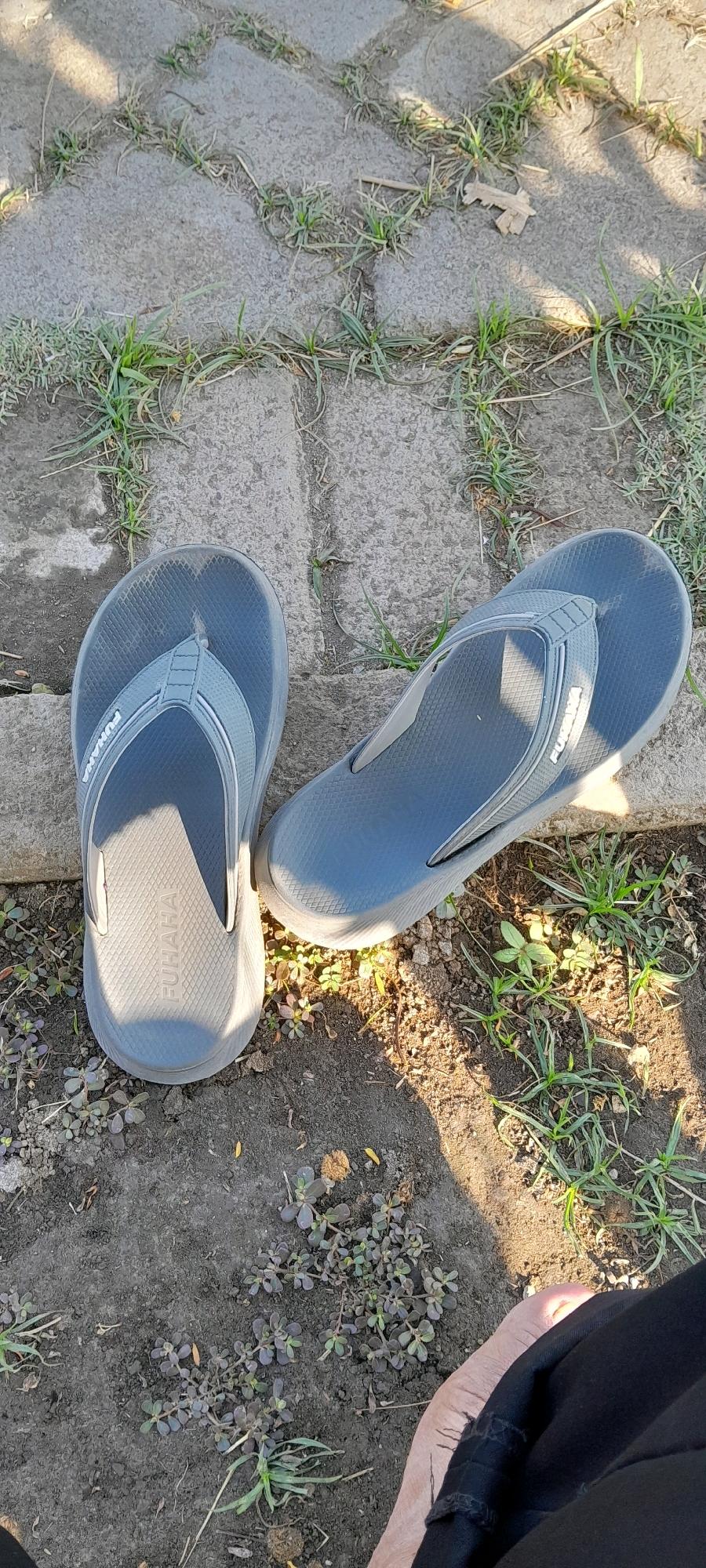 The image shows a pair of gray flip-flops lying on a ground composed of interlocking concrete pavers and dirt, with sparse green grass and small plants growing in the gaps and surrounding areas. The flip-flops are positioned slightly apart, with the left one closer to the viewer and lying on its side, while the right one is more upright. Both flip-flops appear to be made of rubber or a similar material, with a textured footbed and a single strap that goes between the toes. The left flip-flop has a logo or text embossed on its footbed which reads "FUHAHA". The scene is outdoors, as indicated by the natural lighting and vegetation. The shadows suggest the sun is at an angle, possibly late afternoon. There is a hint of a person's foot and black clothing in the bottom right corner of the image. The environment suggests a casual outdoor setting, potentially a pathway or garden. The provided location information, "Bendungan, Indonesia," places this scene within that specific geographical context.