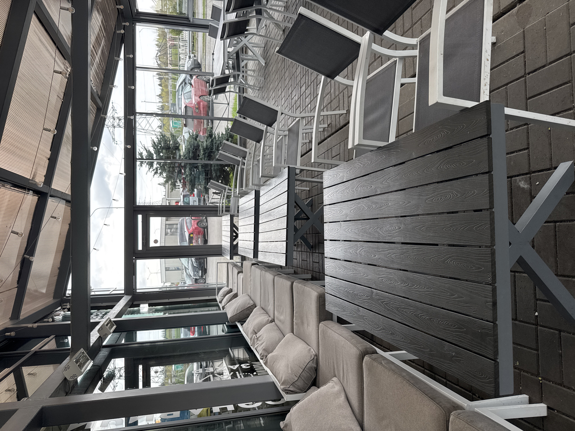 The image depicts an outdoor seating area, likely a patio or cafe, potentially located in or around Medovoye, Russia. The setting includes tables and chairs with white frames and dark gray seats, arranged next to a long, upholstered bench with numerous cushions. The flooring is made of gray paving stones. The area is enclosed by a glass structure supported by a metal frame. Through the glass, a view of the outdoors is visible, including vehicles, trees, and what appears to be power lines. The overall scene presents a modern, outdoor dining or relaxation space. No people are directly visible in the image.