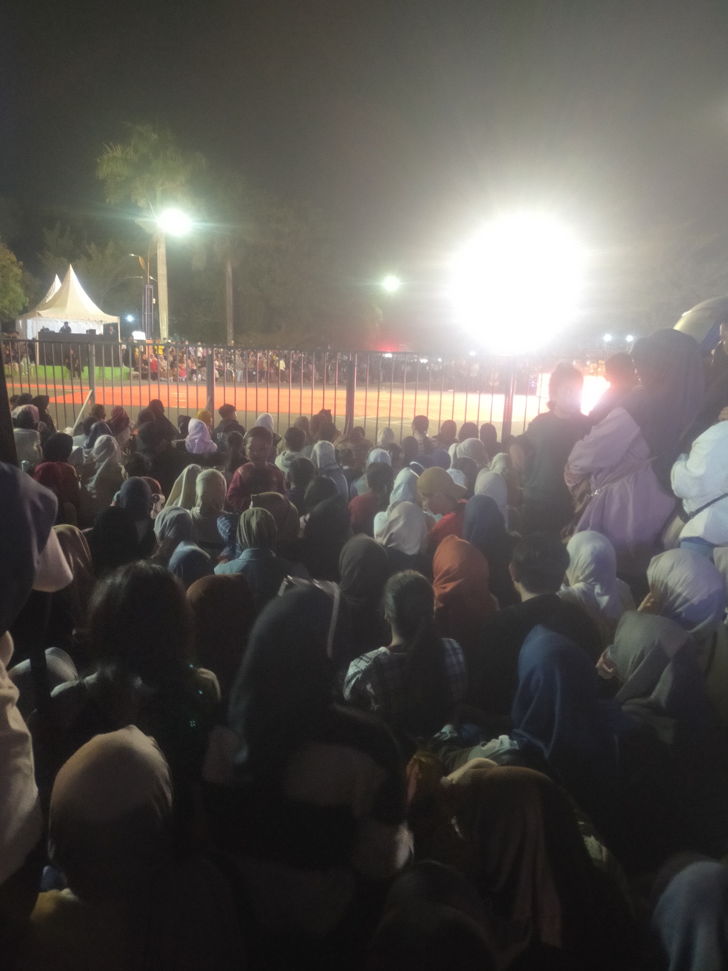 The image depicts a large crowd of people gathered outdoors at night. The foreground is filled with the backs of many individuals, most of whom appear to be wearing headscarves or hijabs. The crowd is densely packed, suggesting a significant event or gathering. In the midground, a metal fence separates the crowd from an area with a white tent structure and palm trees. Beyond the fence, a bright light source illuminates the scene, creating a strong glare and obscuring some details. The overall atmosphere is dark, with artificial lighting contributing to the visibility. The location is identified as Slawi, Indonesia.