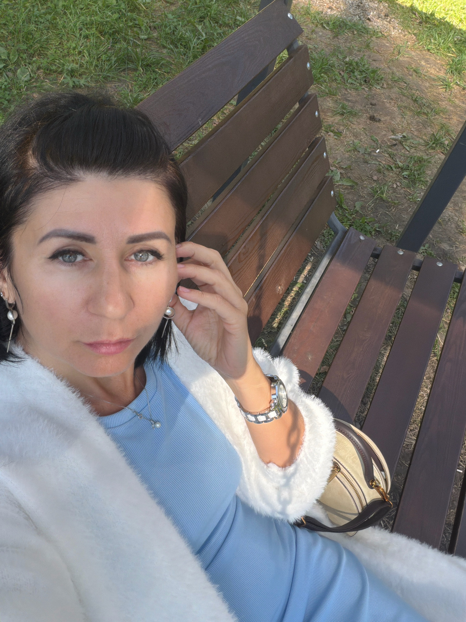 A woman is sitting on a wooden park bench in Angarsk, Russia, taking a selfie. She has dark hair and is wearing a light blue ribbed top, a white fluffy cardigan, and a silver necklace with a pearl pendant. She is also wearing dangling earrings and a silver watch on her left wrist. A light-colored handbag with dark brown trim rests on the bench next to her. The bench is made of dark brown wood slats and has dark metal legs. In the background, there is green grass and dry dirt. The sunlight casts shadows across the scene, indicating it is daytime.