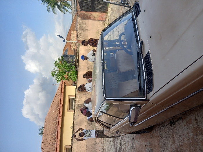 The image shows a brown sedan parked in front of a two-story building. There are six people standing around the vehicle. One person is holding a blue umbrella and another is holding a camera. The house is in the background with a white roof and a brown wall. There are trees and a water tank behind the building. The image was taken in Ita Oniyan, Nigeria, located at the coordinates (latitude: 7.286, longitude: 5.16).