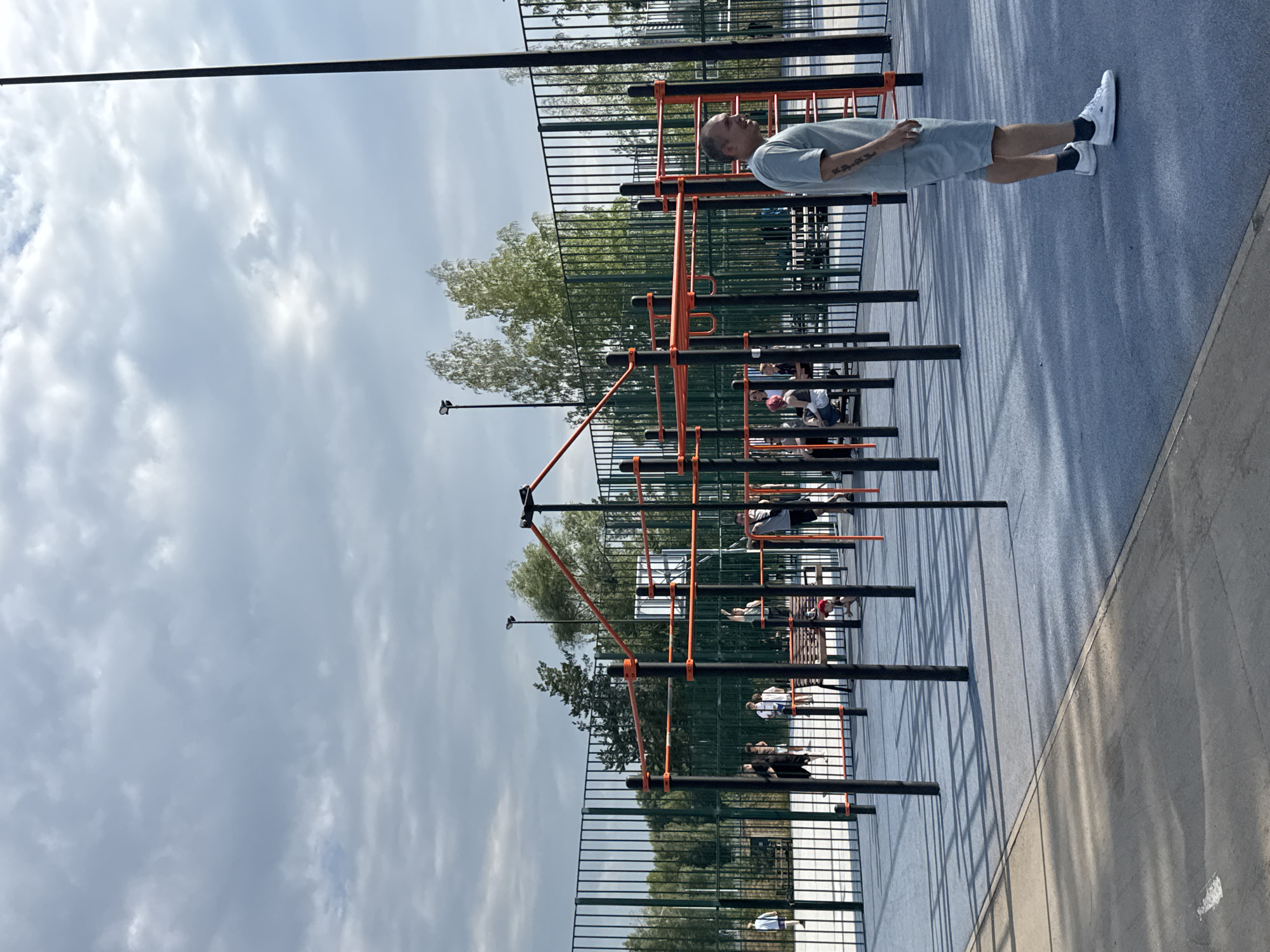The image captures an outdoor sports ground with various exercise equipment. In the foreground, a man in a light blue t-shirt and shorts stands facing right, looking away from the camera. He is wearing white sneakers and dark socks. Behind him, a complex structure of black poles and orange bars, typical of a calisthenics park, dominates the scene. Several other individuals are scattered throughout the sports ground, some on benches, others appearing to engage in activities. A green fence encloses the area, with trees and sky visible beyond. The ground is a smooth, light blue surface, with darker blue sections and concrete pathways. The lighting suggests it is daytime with overcast skies, indicated by the soft light and scattered clouds in the sky. The overall impression is of a public recreational space. The location is identified as Novosibirsk, Russia.