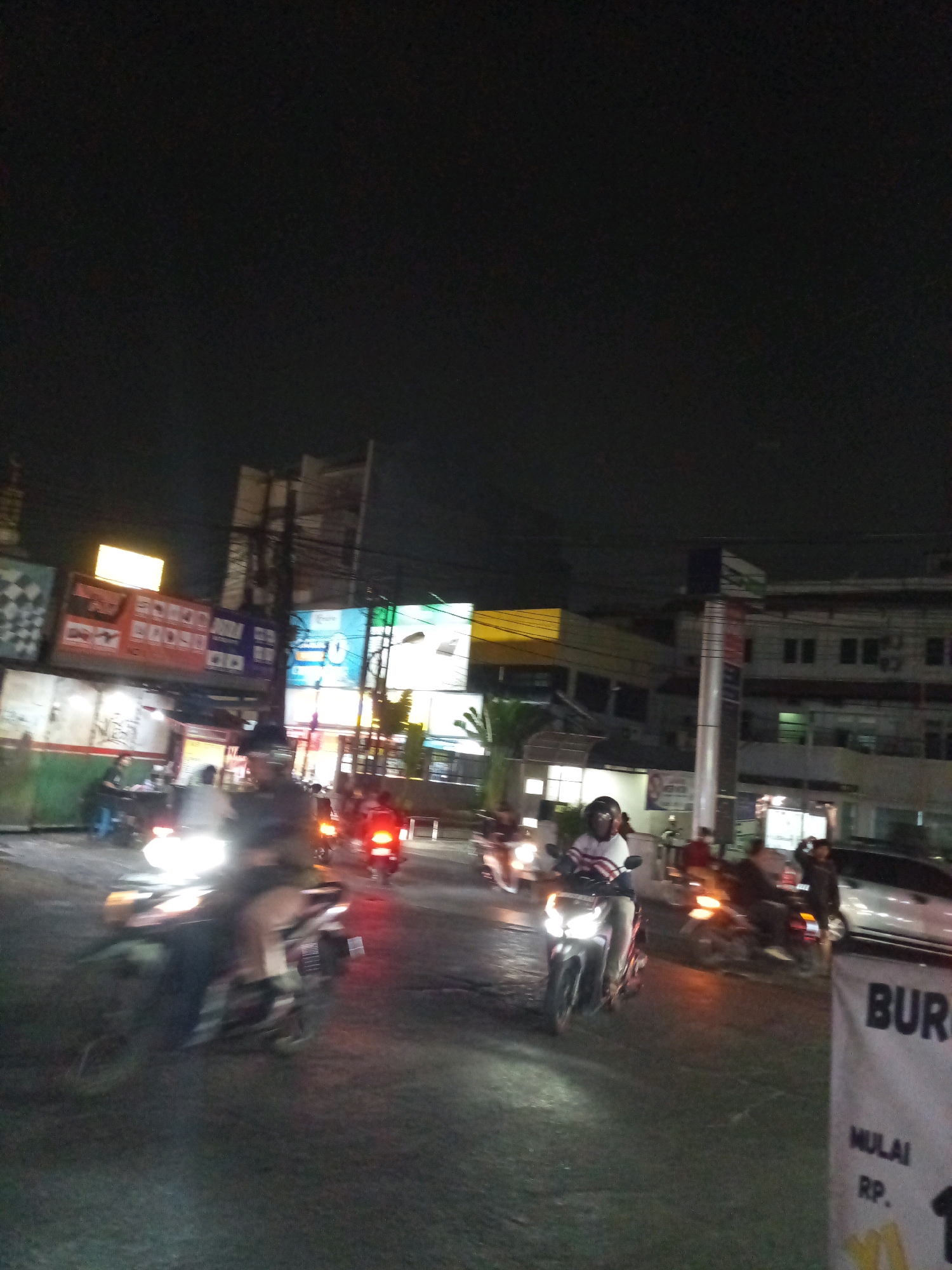 The image depicts a nighttime street scene in Bekasi, Indonesia, characterized by a dark sky and illuminated by artificial lights from buildings, signs, and vehicle headlights. The foreground shows a street with several motorcycles in motion, some with their headlights on, creating blurred streaks of light. One motorcycle in the center foreground has its rider clearly visible, wearing a helmet and a striped shirt. In the background, buildings with various signs and lights are visible, some of which appear to be commercial establishments. A tall pole with numerous wires stretches across the upper portion of the scene. A banner with the word "BURG" is visible in the lower right corner. The overall atmosphere is that of a busy urban environment at night.