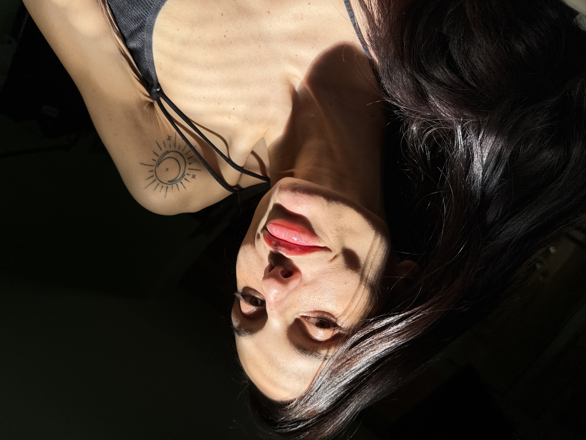 An upward-angled portrait features a female individual with long, dark brown hair, positioned against a deeply shadowed background. Her face, partially illuminated by a strong, directional light source from the upper left, shows a smooth complexion. Her eyes are open and directed towards the viewer. Her lips are full and glossy, coated with a reddish-pink substance. A prominent tattoo depicting a crescent moon encircled by a sun with radiating lines and two small stars is visible on her left upper arm. She is wearing a dark-colored top with thin spaghetti straps. Shadows sharply define her facial features and upper torso, contrasting with the illuminated areas. The environment appears to be an interior setting, but no specific architectural or furnishing details are discernible to identify a precise location within Pleven, Bulgaria, due to the minimal lighting and featureless background.