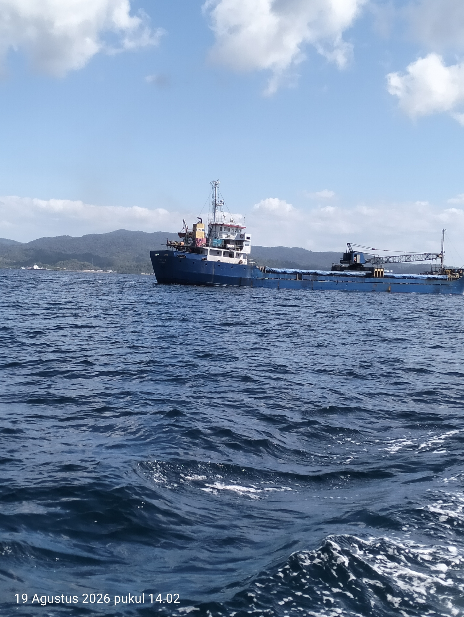 A wide shot captures a large, blue cargo ship navigating the deep blue sea under a bright sky dotted with white clouds. The ship, with its white superstructure and a prominent crane structure, is positioned in the mid-ground, heading towards the right of the frame. The foreground is dominated by the choppy texture of the sea. In the background, a chain of lush green hills forms a natural coastline, suggesting a tropical or subtropical environment. The timestamp "19 Agustus 2026 pukul 14.02" is overlaid on the bottom left of the image, indicating it was taken on August 19, 2026, at 2:02 PM. The location is specified as Kendari, Indonesia.