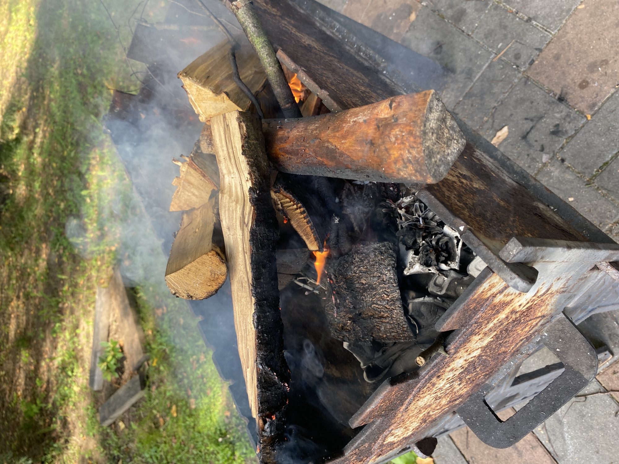 The image depicts a fire pit filled with burning logs and emanating smoke. The fire pit is made of rusted metal and is situated on a paved surface, likely a patio or backyard. In the background, there is a grassy area with some fallen leaves and dappled sunlight, suggesting an outdoor setting. The scene appears to be in an urban or suburban environment, possibly a garden or park within the city of Зимородок Днп, Russia. There are no people visible in the image, and the focus is on the burning fire and its immediate surroundings.