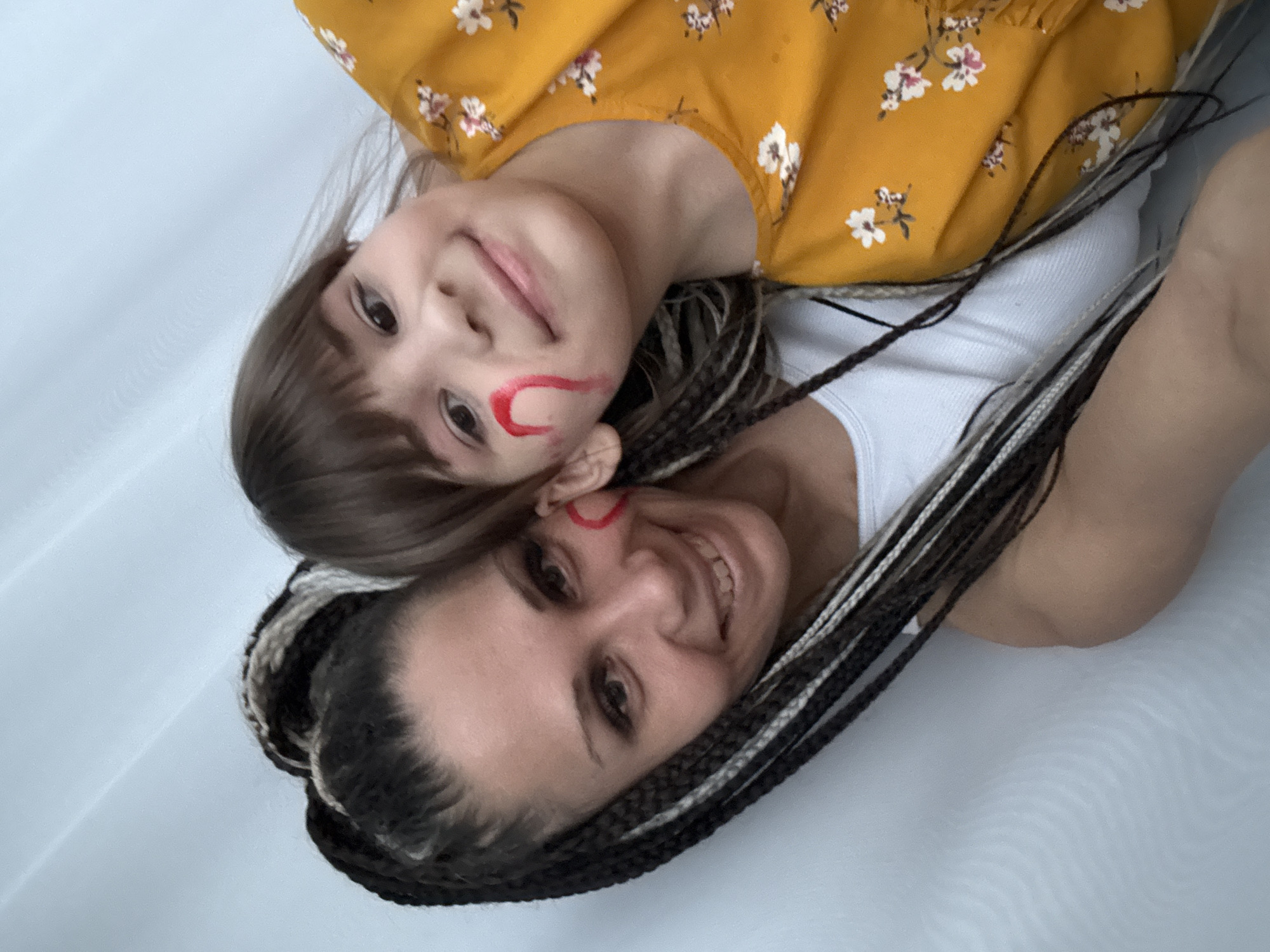This image, taken in Rusanovo, Russia, features two females, likely a mother and daughter, lying down on a plain, light-colored surface, possibly a bed or blanket. The photograph is taken from above, looking down on the subjects.
The adult woman, positioned closer to the bottom of the frame, has dark hair styled in numerous dreadlocks or braids, interspersed with some lighter strands. She is wearing a white ribbed tank top. A small red, crescent-like marking is visible on her right cheek, near her ear. She is smiling warmly and looking towards the camera.
Resting her head above the woman's, the young girl has brown hair and is wearing a mustard yellow top with white and pink floral patterns. A more pronounced red crescent shape is painted on her right cheek. She also smiles sweetly, looking directly at the camera.
The scene conveys a sense of joy and affection between the two individuals, possibly captured during a playful moment indoors. The lighting is bright and even, indicating a daytime setting. There is no visible text in the image.