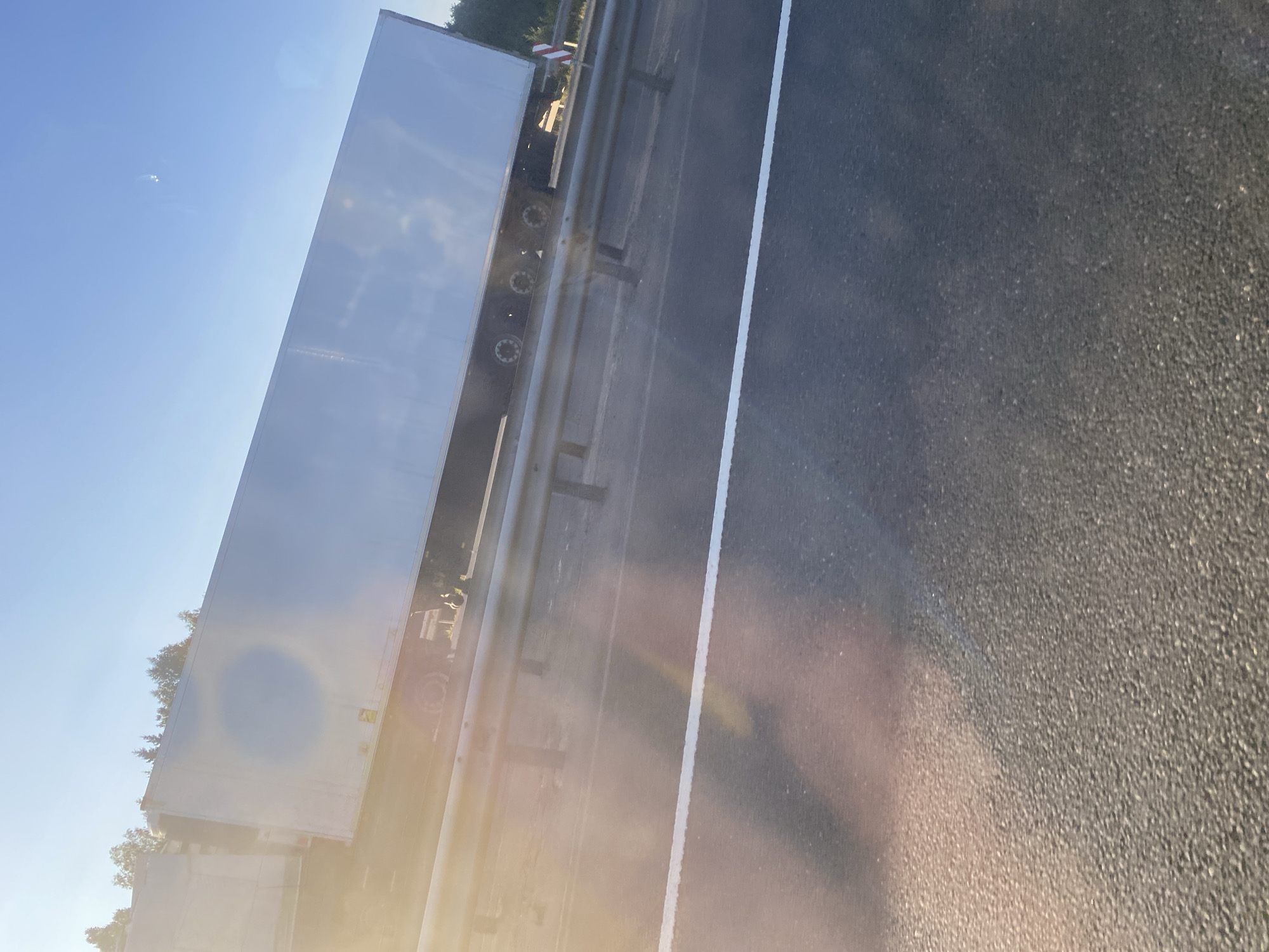 An exterior, eye-level shot captures a tilted white semi-trailer truck, its metallic surface reflecting the bright, hazy sun. The truck is positioned on a roadway, partially obscured by a silver guardrail. The sky above is a clear, pale blue, with a few faint white clouds in the distance. A bright white line runs horizontally across the asphalt in the foreground. Trees form a dark green backdrop to the right of the truck. The overall impression is one of a sunny day on a highway, with the glare of the sun creating a lens flare effect across the image.