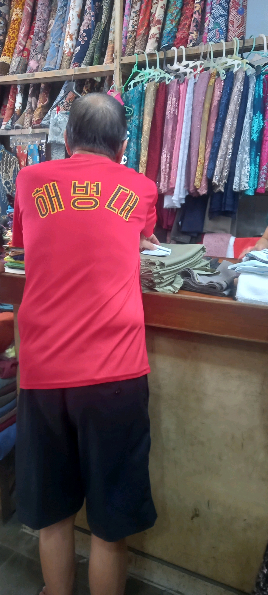 In Jember, Indonesia, a person wearing a red t-shirt with Korean text and dark shorts stands in front of a shop displaying various colorful fabrics and textiles. The person's back is to the camera, and they are interacting with fabrics that are folded on a wooden counter. The shop appears to be selling fabrics, possibly for clothing or crafts, as indicated by the racks of material and the way the fabrics are presented. The background shows shelves laden with rolls of patterned fabric, suggesting a retail environment focused on textiles. The lighting is somewhat dim, casting a naturalistic feel to the scene.