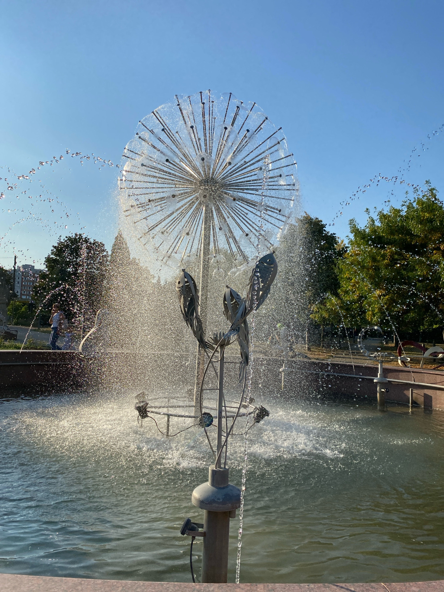 This is a photograph of a modern dandelion-shaped fountain in Chișinău, Moldova. The fountain's structure is made of metal rods radiating from a central point, with water spraying from the ends of these rods, creating a spherical effect. Below the dandelion structure are two stylized metal fish sculptures. The fountain is situated in a pool of water, surrounded by a stone border. In the background, there are trees, buildings, a clear blue sky, and a person walking near the fountain. The lighting suggests it is daytime.