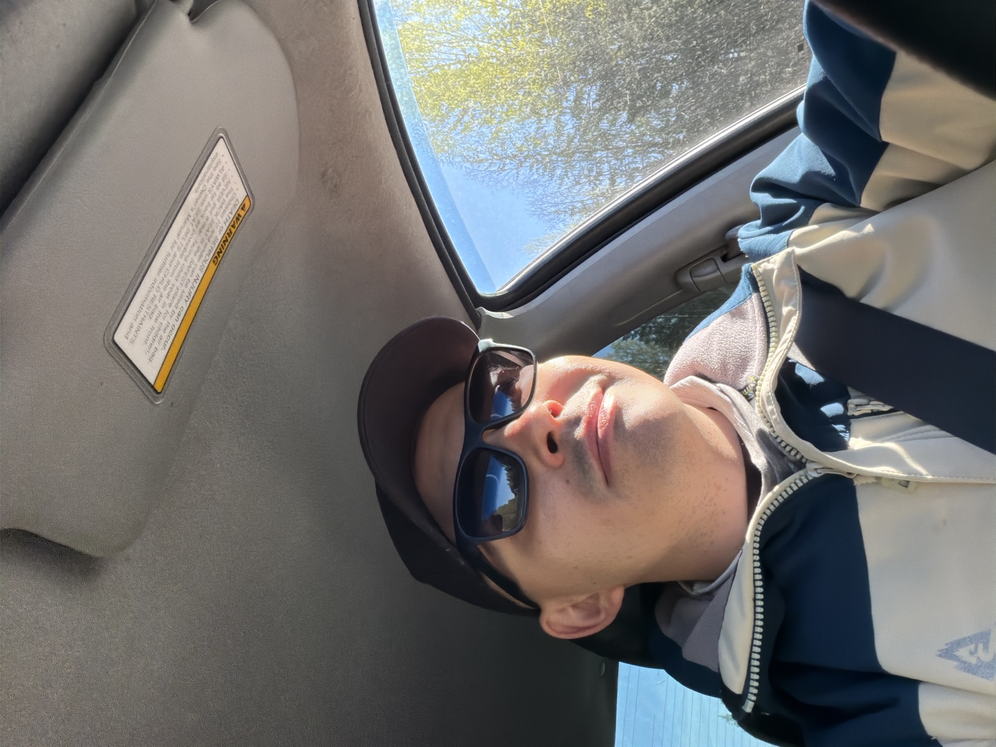 The image is an upward-angled shot taken from inside a car, presumably in motion or at a standstill. A person is visible, wearing a dark baseball cap and sunglasses, with a jacket in shades of blue and white. The car features a sunroof and a sun visor with a visible warning label. Outside, the sky is visible through the sunroof, along with trees. Given the language of the warning label, this is unlikely to be in Russia, and is thus not located in Severnyy, Russia. The setting seems to be during daylight hours with bright sunlight.