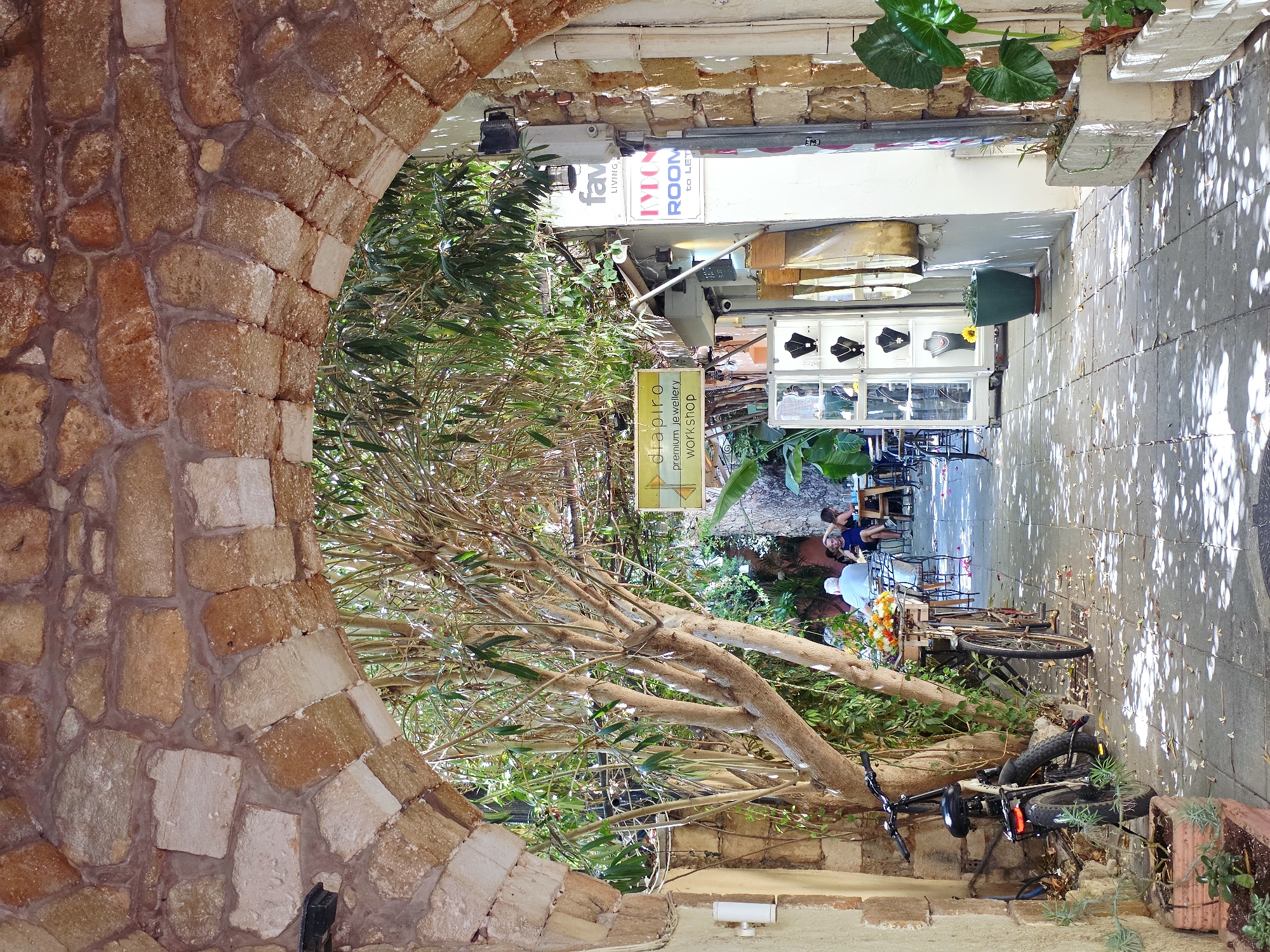 The image depicts a sun-dappled stone archway leading into a narrow, shaded alleyway in Chania, Greece. The arch is constructed from rough-hewn, reddish-brown and cream-colored stones. Beyond the arch, a leafy tree with a thick trunk is prominent, its branches extending overhead and casting shadows on the cobblestone path below.
To the left, a textured, pale yellow wall forms the side of the alley, with a white outdoor light fixture mounted on it. A bicycle with thick tires and a black seat is parked against this wall, partially obscured by a potted plant. Further down, another bicycle with a basket filled with colorful flowers is visible.
On the right side of the alley, a white building facade is visible with several signs. One sign, in yellow, reads "diapiro premium jewellery workshop". Below it, another sign in blue and white reads "KIMON ROOM to LET". A shop window displays jewelry on mannequins.
Several people are seated at tables in the background, suggesting an outdoor cafe or restaurant. One person is wearing a white shirt, another a dark dress. The atmosphere appears relaxed and summery. Potted plants and planters are strategically placed along the path, adding greenery to the scene. The ground is paved with grey cobblestones, scattered with fallen leaves and petals. The sunlight filters through the leaves, creating a dappled effect on the path.