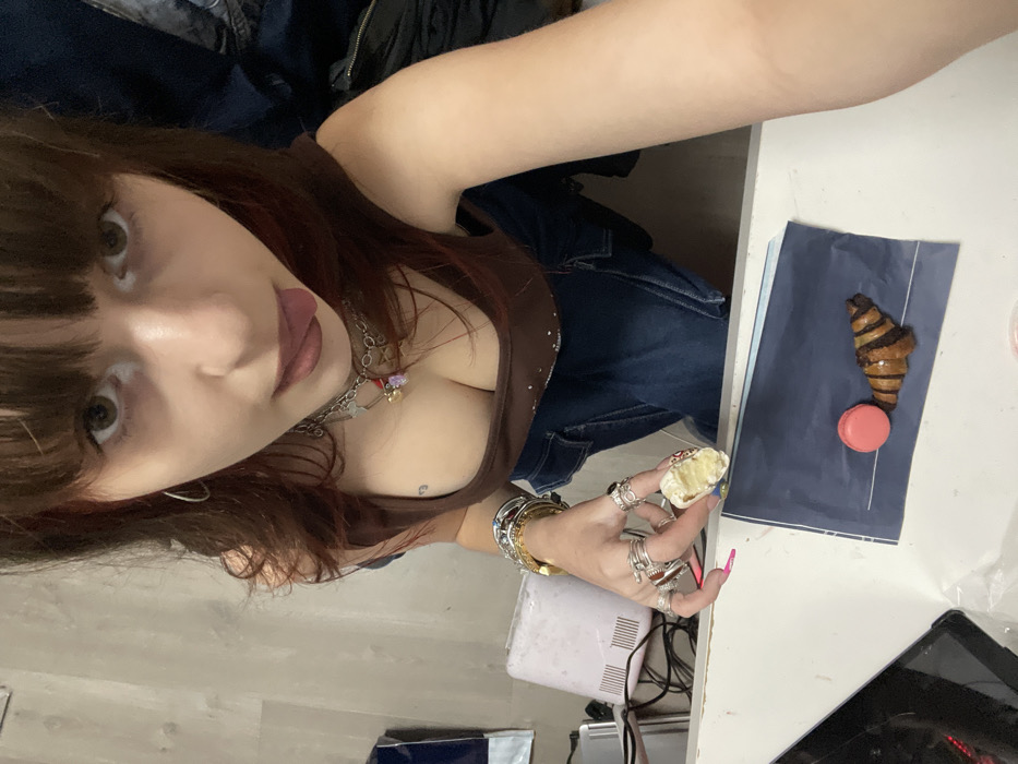 A young woman with dark hair and bangs poses in a selfie, sticking her tongue out slightly. She wears a brown tank top and multiple silver necklaces. Bracelets and rings adorn her hand, which holds a pastry with cream filling. A table features a pink macaroon, a chocolate croissant on a blue napkin, a nail lamp, and a tablet. She is wearing jeans.