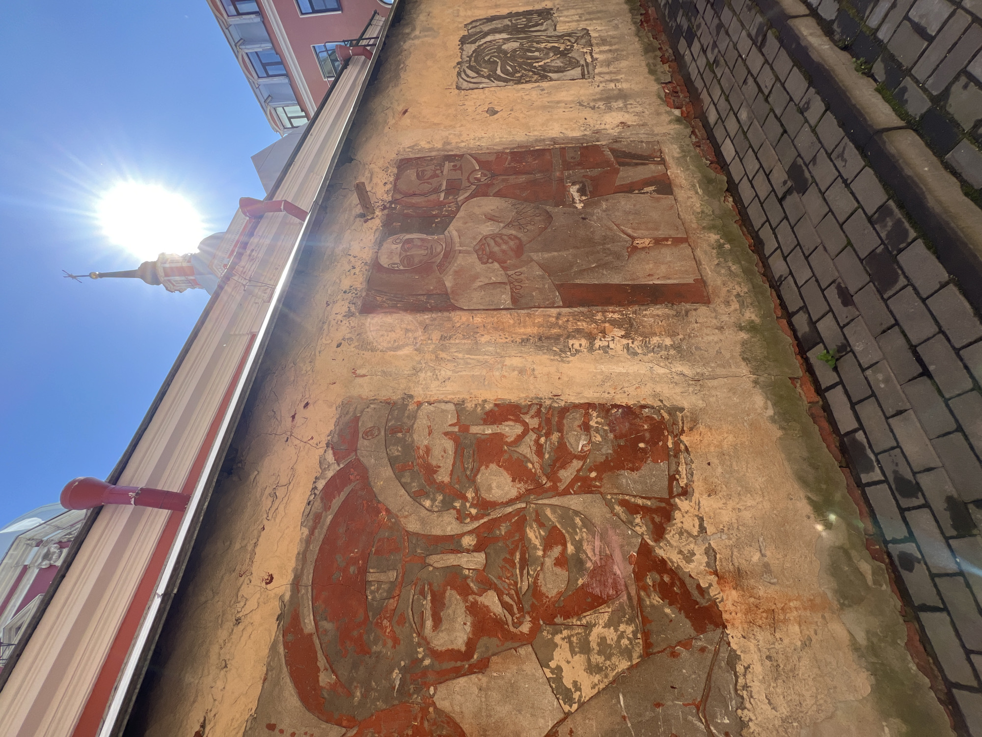 The image depicts a dilapidated wall in Saint Petersburg, Russia, featuring three faded murals or reliefs. The top mural is abstract and indistinct. The middle mural shows two figures, possibly military or political figures, standing closely together. The bottom mural is a close-up of a face wearing what appears to be a helmet or headdress. The wall is adjacent to a brick-paved walkway. The top of an adjacent building with a spire is visible, suggesting the image was taken near a historical or architectural landmark. The sun is bright in the sky, casting harsh shadows.