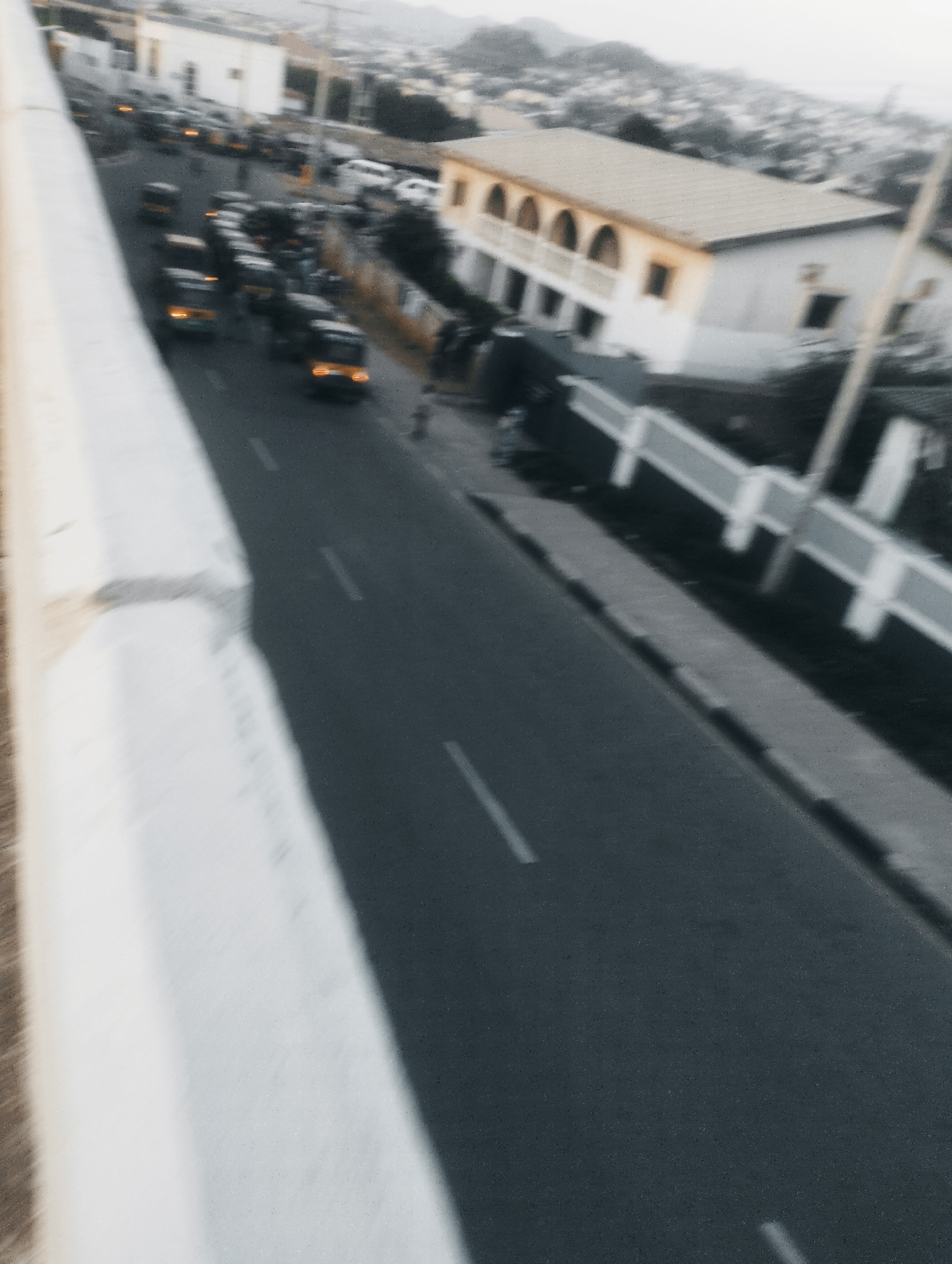 The image captures a view of a street in Jos, Nigeria, from an elevated perspective, possibly a bridge or overpass. The foreground is dominated by a thick, white concrete barrier, with a blurred texture suggesting motion or a close-up, out-of-focus shot. The street itself, paved with dark asphalt, is visible below, with faint white lane markings.
Several vehicles, including what appear to be minibuses or commercial vans, are traveling on the road. Some of these vehicles have their headlights or taillights illuminated, suggesting it might be dusk or an overcast day. The traffic seems to be moving, with a noticeable congestion or slow-down in some sections. In the background, a residential area with buildings and houses can be seen, set against a backdrop of hills. One building in particular stands out with its arched windows and a light-colored facade, possibly a public building or a large house. The overall atmosphere appears to be that of a typical urban scene in Nigeria, with the characteristic vehicles and architecture. The lighting suggests it is late afternoon or early evening. There is no visible text in the image.