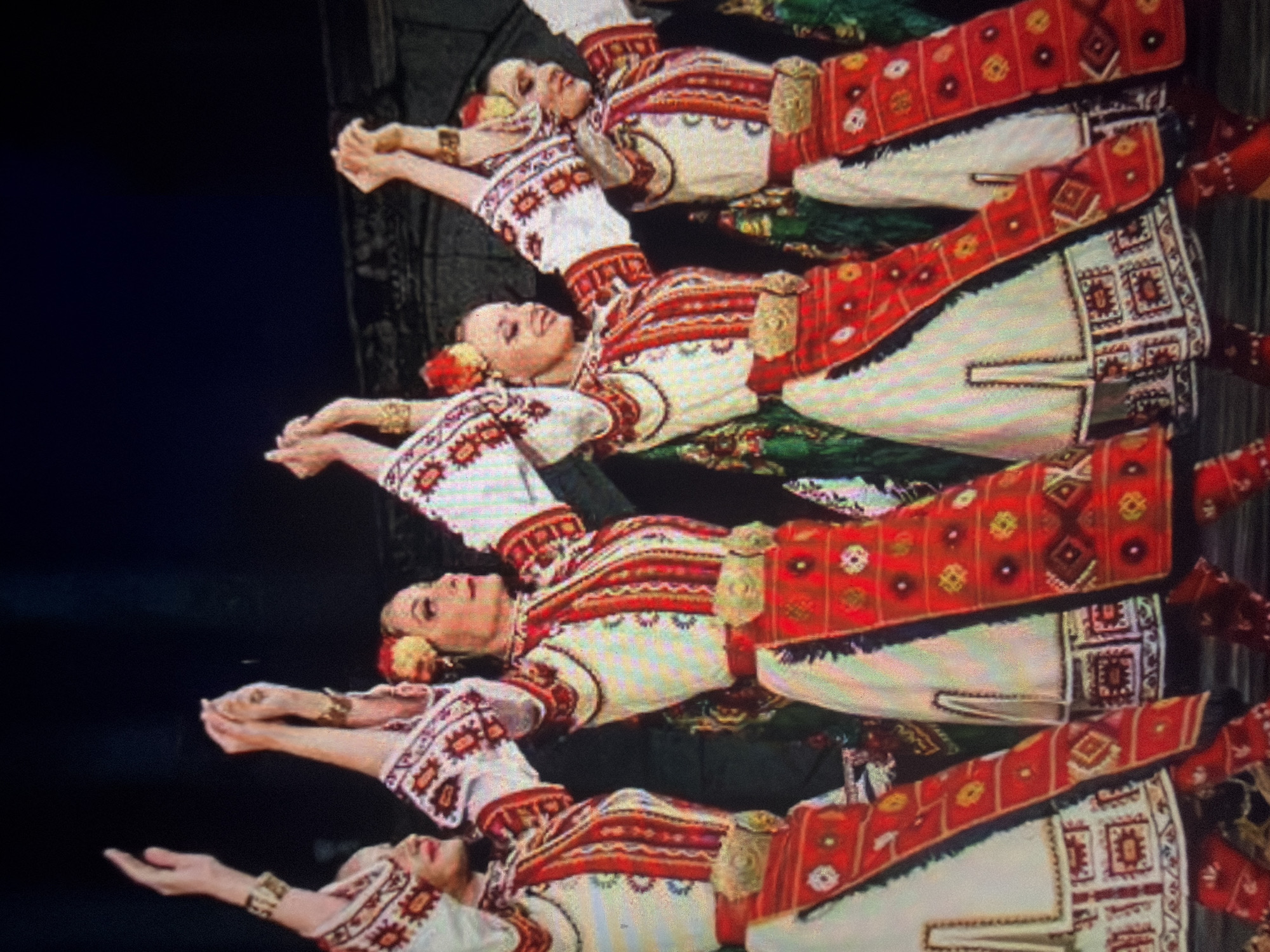 Four women are depicted in a choreographed line, performing a traditional dance in folk costumes. The scene is located in the city of Shumen, Bulgaria. Each woman wears a long-sleeved white garment featuring intricate red, black, and geometric embroidery patterns on the sleeves and bodice. A prominent wide, ornate gold-colored metal belt with a large central buckle cinches the waist. Over the white garment, a rectangular red fabric panel, heavily embroidered with multi-colored geometric designs, is visible on the front. Gold-colored bracelets adorn their wrists. Each dancer's hair is ornamented with red and yellow flowers positioned above the right ear. Their arms are extended upwards, with hands interlinked, forming a continuous chain above their heads. Their facial expressions convey serene focus or slight smiles. The background is dark, consistent with a stage or performance setting, with minimal structural elements faintly discernible and areas of dark green fabric visible behind the lower half of the central figures. Red, embroidered footwear is observable on the dancer positioned furthest to the right.