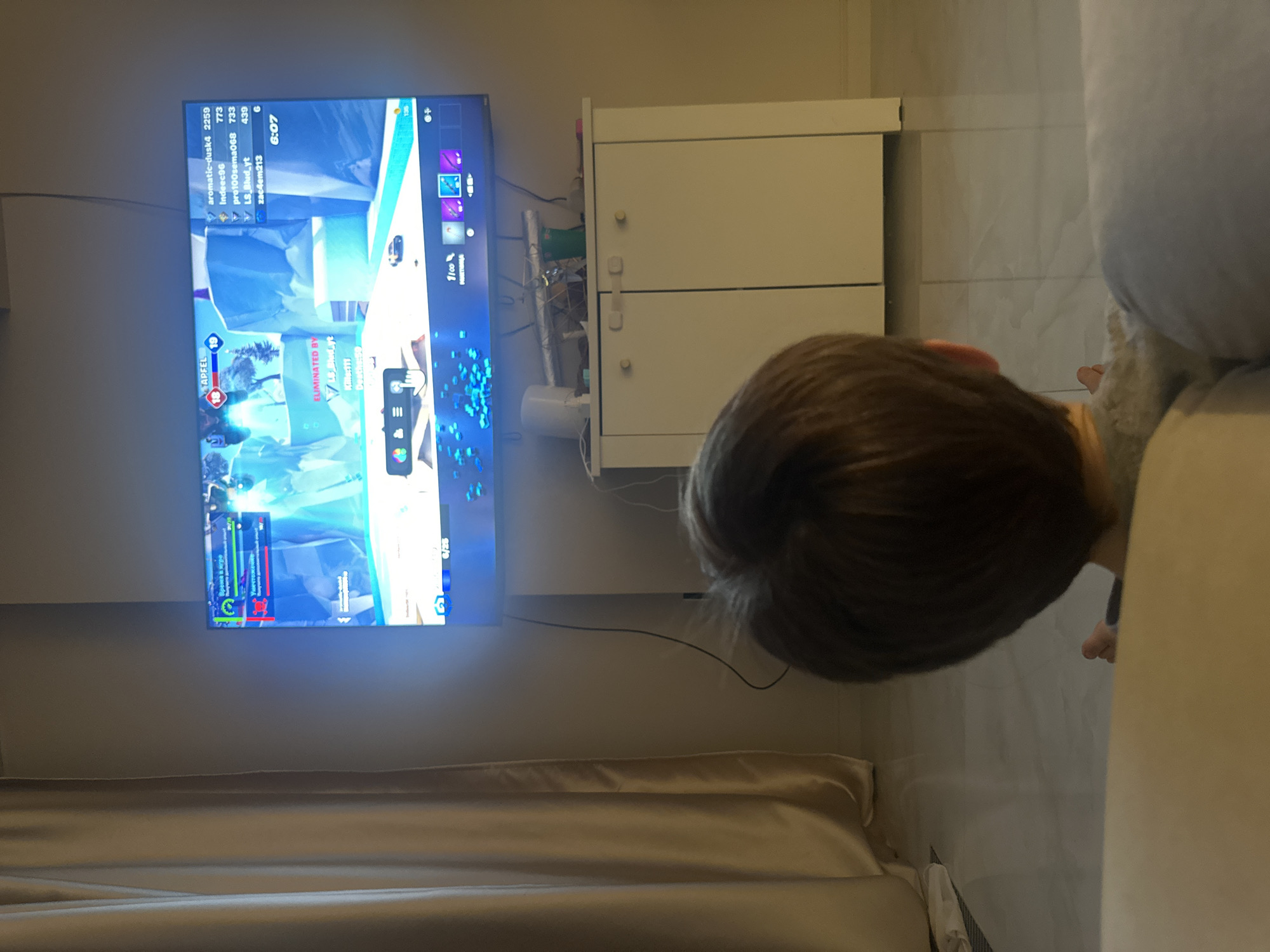 The image shows a child with brown hair watching a video game on a large television. The game is a first-person shooter with a futuristic or fantasy setting. The child is positioned in front of a white cabinet and a light beige couch. The game displays player information like "APFEL", "ELIMINATED BY LB Blud_yt", along with the time "6:07". The location seems to be indoors, potentially a living room, with beige curtains. The image does not contain the information about Khimki, Russia.