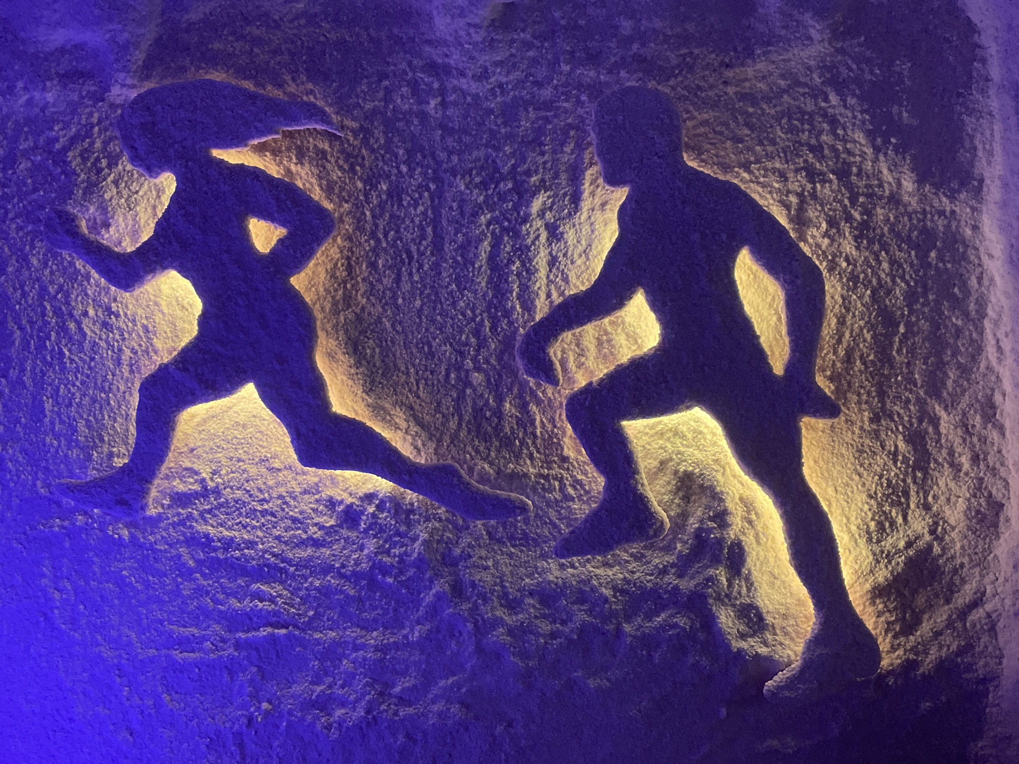 The image shows a salt cave in Vinnytsia, Ukraine. The cave has carvings of a woman and a man, both depicted in running poses, illuminated from behind. The wall surrounding the carvings is textured and appears to be made of salt. The lighting is primarily blue on the left and lower areas, shifting to yellow-orange behind the figures.