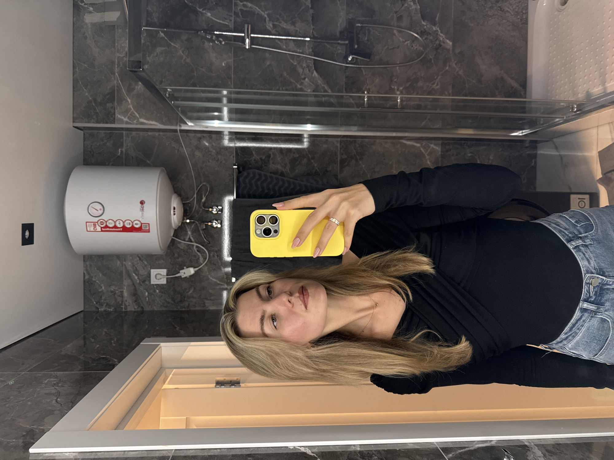 A woman in a black top and jeans is taking a selfie in a bathroom. The bathroom has a gray marbled wall and floor. There's a water heater mounted on the wall to the left of the woman, along with a showerhead and glass enclosure above. The bathroom is located in Unknown, Unknown.