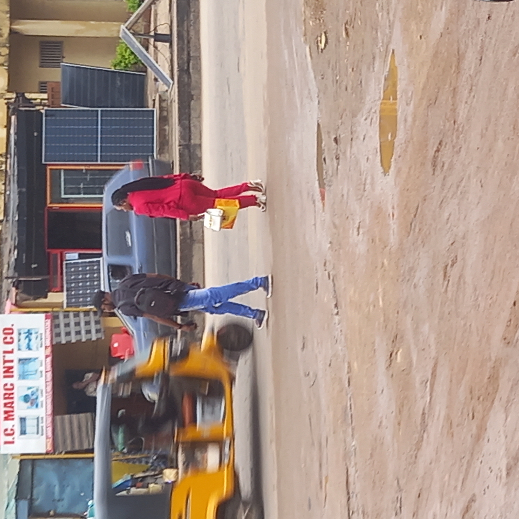 The scene appears to be a street in Dadi Riba, Nigeria.
A yellow motorized tricycle ("keke") is in the lower left portion of the frame, positioned in the street, with a person visible inside. In front of the tricycle, a man wearing a black shirt, blue jeans, and carrying a backpack is crossing the street. Further back, a woman dressed in a red outfit is also crossing the street, carrying a light-colored bag.
Behind the woman and man is a blue sedan parked on the side of the road. Next to the blue car, a building is visible with solar panels on its front. A sign for "I.C. MARC INTL CO." is displayed on the building's facade.
The road itself is made of a light-brown surface and shows signs of wear, with puddles or damp patches visible.
