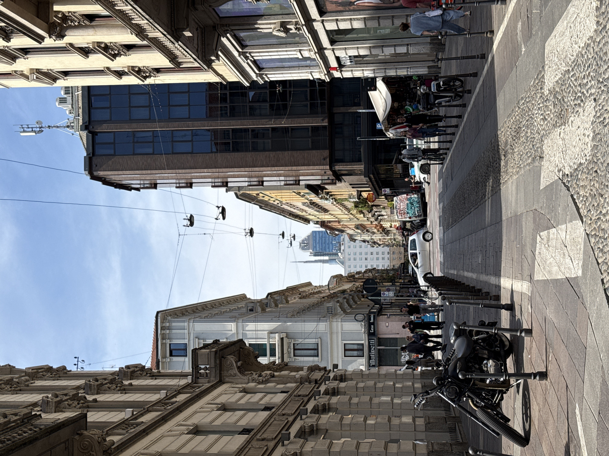 The image captures a street scene in Milan, Italy, characterized by narrow roads flanked by buildings with ornate facades. In the distance, the Duomo di Milano is visible. Pedestrians stroll along the street, and a motorcycle is parked along the curb. Overhead, power lines stretch across the street, connecting the buildings. The sky is clear and blue.