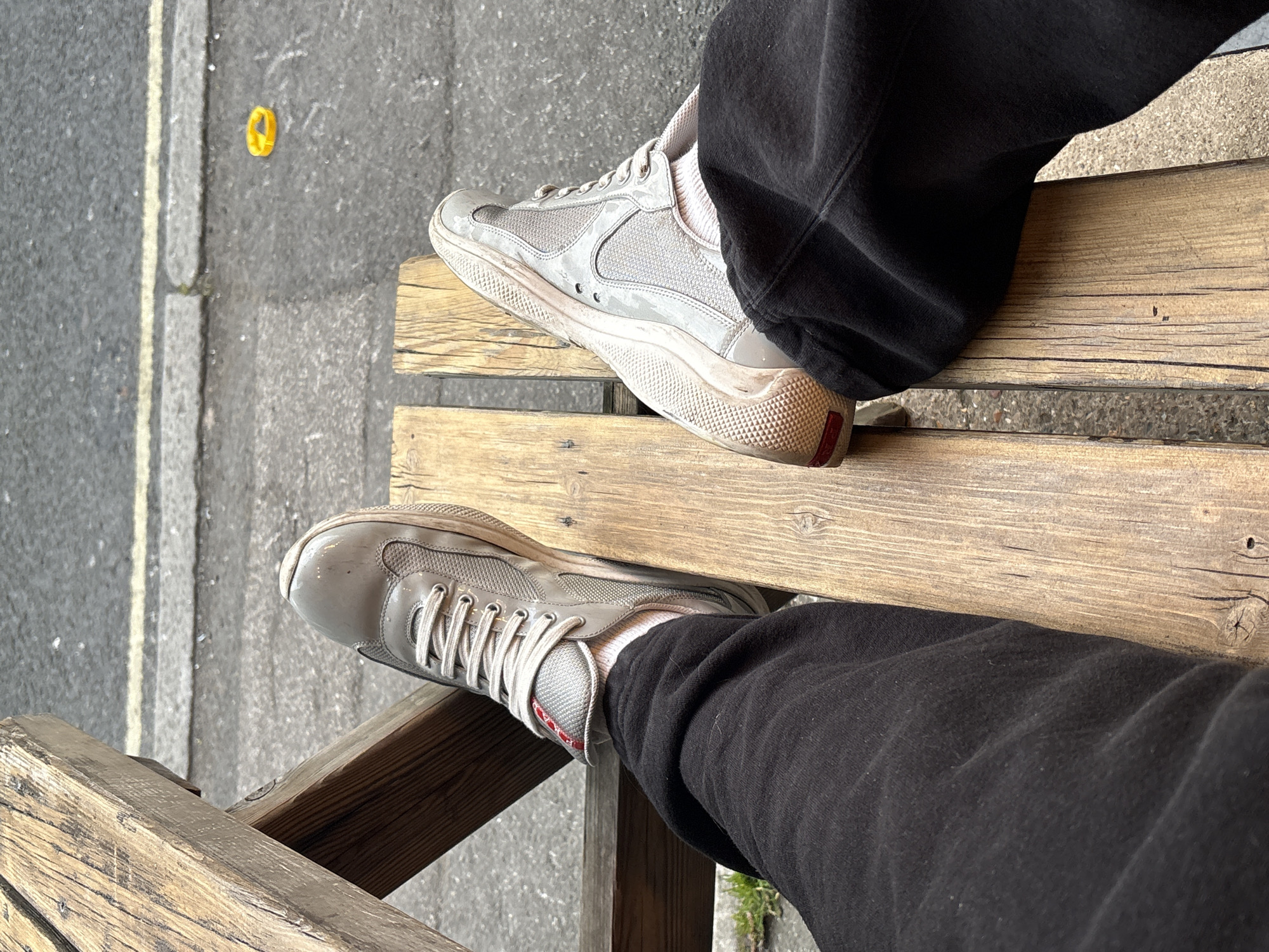 A first-person or overhead shot captures the lower legs and feet of a single individual. The person wears black, relaxed-fit trousers and white ankle-length socks. Both feet are clad in light grey Prada America's Cup sneakers, which exhibit signs of wear and dirt, with subtle red accents visible on the left shoe's tongue and the heels of both. The feet rest upon a weathered, light brown wooden structure, likely an outdoor bench or pallet. Beyond this structure, a grey asphalt road is visible, featuring a yellow painted line and a grey concrete curb. A small, bright yellow, circular object lies on the asphalt in the mid-ground to the right. The scene is illuminated by natural daylight. The depicted urban street environment is characteristic of the city of London, United Kingdom, though a specific location within the city cannot be identified from the imagery.