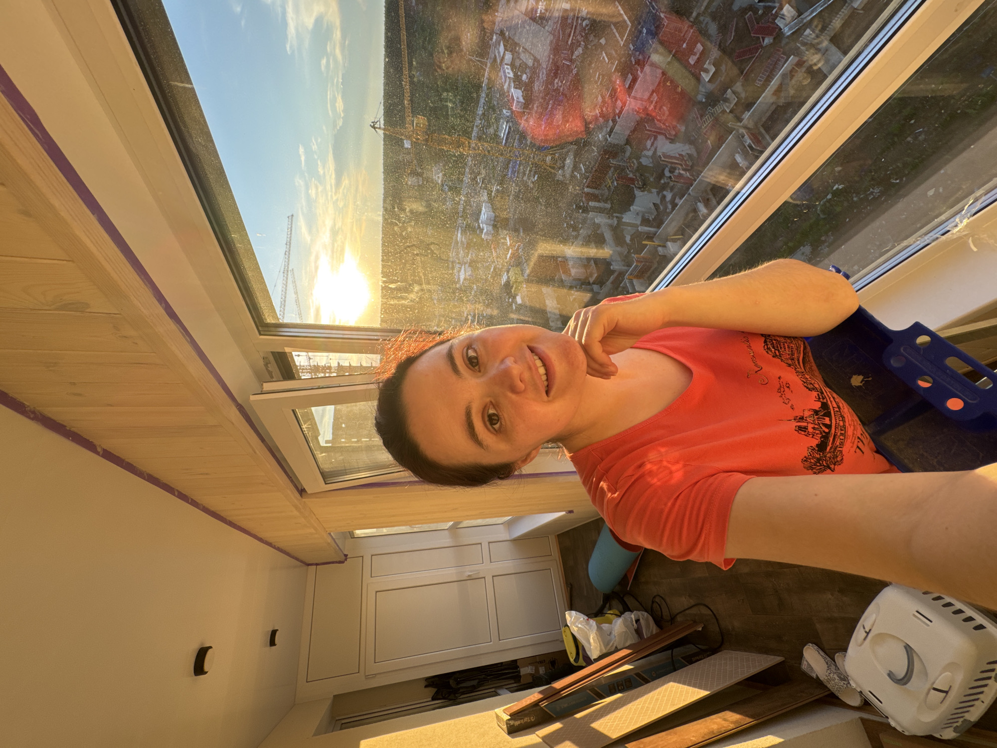 A young woman is taking a selfie on a balcony overlooking a construction site at sunset in Izhevsk, Russia. The woman is smiling and leaning her chin on her hand. She is wearing an orange t-shirt with a graphic of Notre Dame Cathedral. Behind her, the balcony is cluttered with building materials, including wood planks, a blue yoga mat, and a white pet carrier. The large window shows a construction site with cranes and unfinished buildings, bathed in the warm glow of the setting sun. The sky is clear with a few clouds.