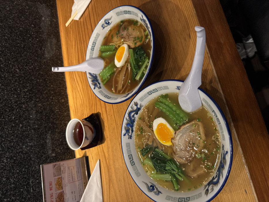 The scene depicts an overhead view of two bowls of ramen served on a light-colored wooden table inside a restaurant in Bangkok, Thailand. Each white bowl features a blue dragon motif and a geometric border, containing a dark broth with noodles, sliced pork, a halved soft-boiled egg with a bright orange yolk, green leafy vegetables, bamboo shoots, and a garnish of white sesame seeds and chopped green onions. A white ceramic or plastic ramen spoon rests in each bowl. To the left, a small, decorative teacup containing a dark beverage sits on a black square saucer. A partially visible menu stand displaying text and images is positioned behind the teacup. A white folded napkin or paper is present near the menu. In the upper right, chopsticks wrapped in white paper are visible. The background consists of a dark, textured wall. No individuals are present.