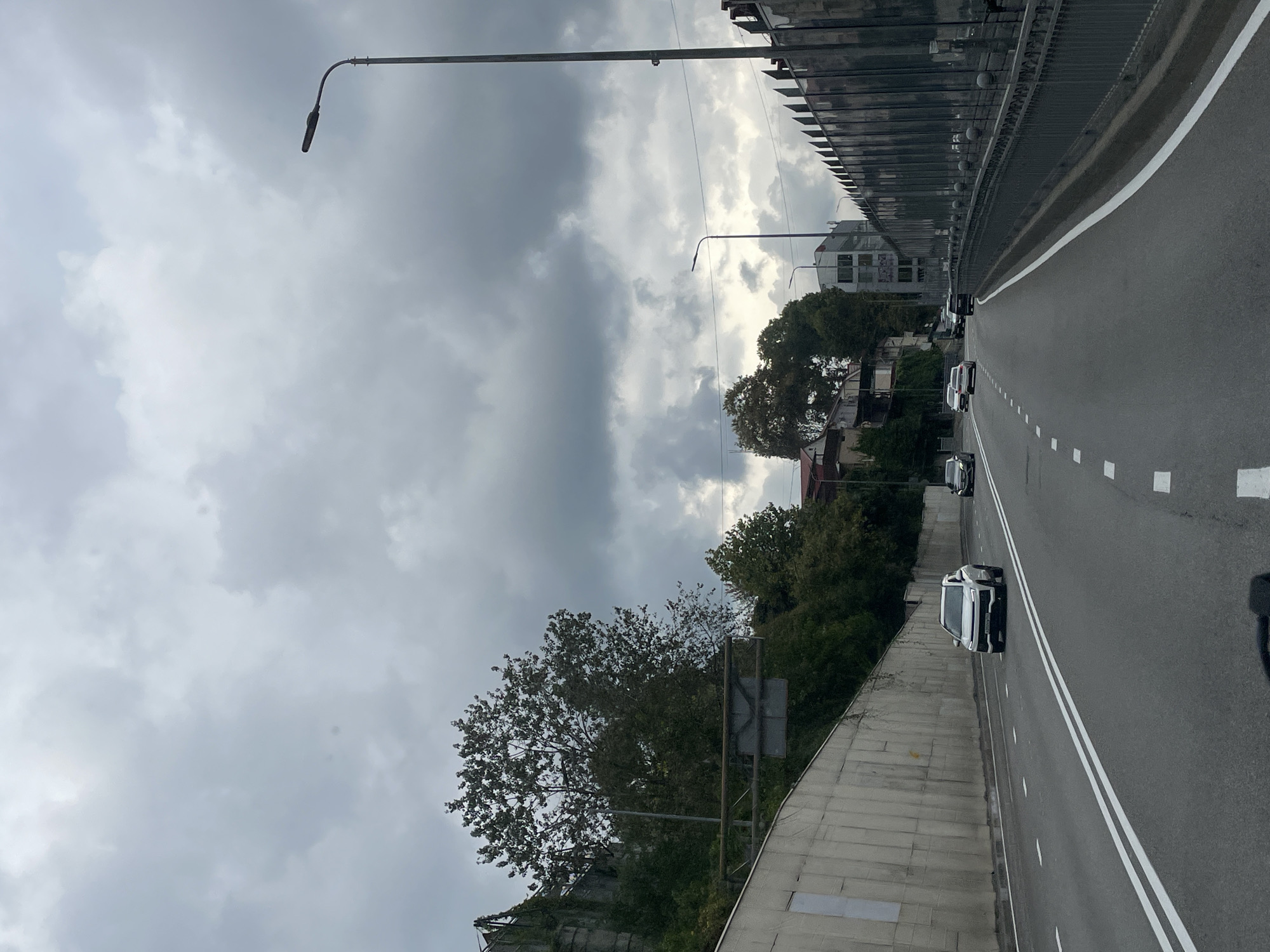 The image depicts a cloudy day in Sochi, Russia. The view is from a vehicle traveling on a multi-lane road, possibly a highway or a major thoroughfare. On the left, a high concrete retaining wall curves along the road, overgrown with some vegetation. Trees and buildings are visible on the slope above the wall. On the right, a metal fence with decorative elements runs alongside the road, separating it from buildings in the background. Several cars are driving on the road, including a white SUV in the foreground and other vehicles further ahead. Streetlights are present along the roadside, and traffic markings are clearly visible on the asphalt. The sky is filled with heavy, gray clouds, suggesting overcast weather. The overall scene is that of a typical urban or suburban road environment under a cloudy sky.