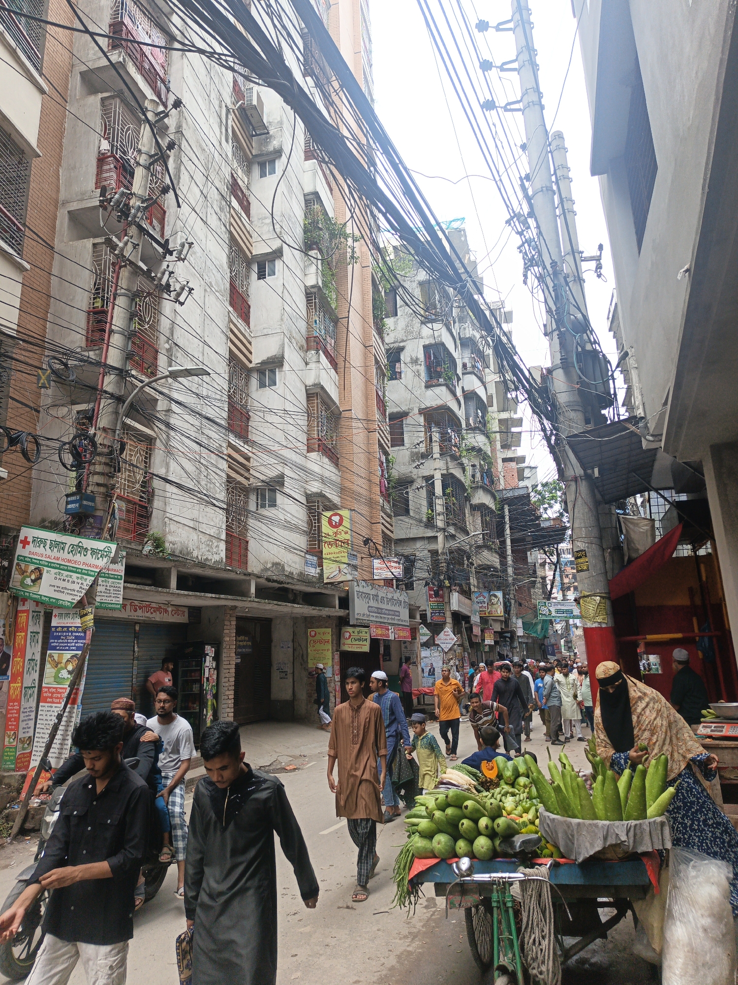 The image depicts a bustling street scene in an urban environment, likely in South Asia. The narrow street is lined with multi-story buildings constructed with concrete and brick, adorned with numerous balconies and exposed electrical wiring that crisscrosses overhead. The architecture suggests a dense urban setting with limited open space. A significant number of people are present, walking, shopping, and interacting. The foreground features a cart laden with fresh produce, primarily green vegetables, being attended to by a woman wearing a niqab. Several individuals are seen browsing the produce. Further down the street, a crowd of men and women are visible, some dressed in traditional attire. The overall atmosphere is one of lively commerce and daily life in a crowded city.