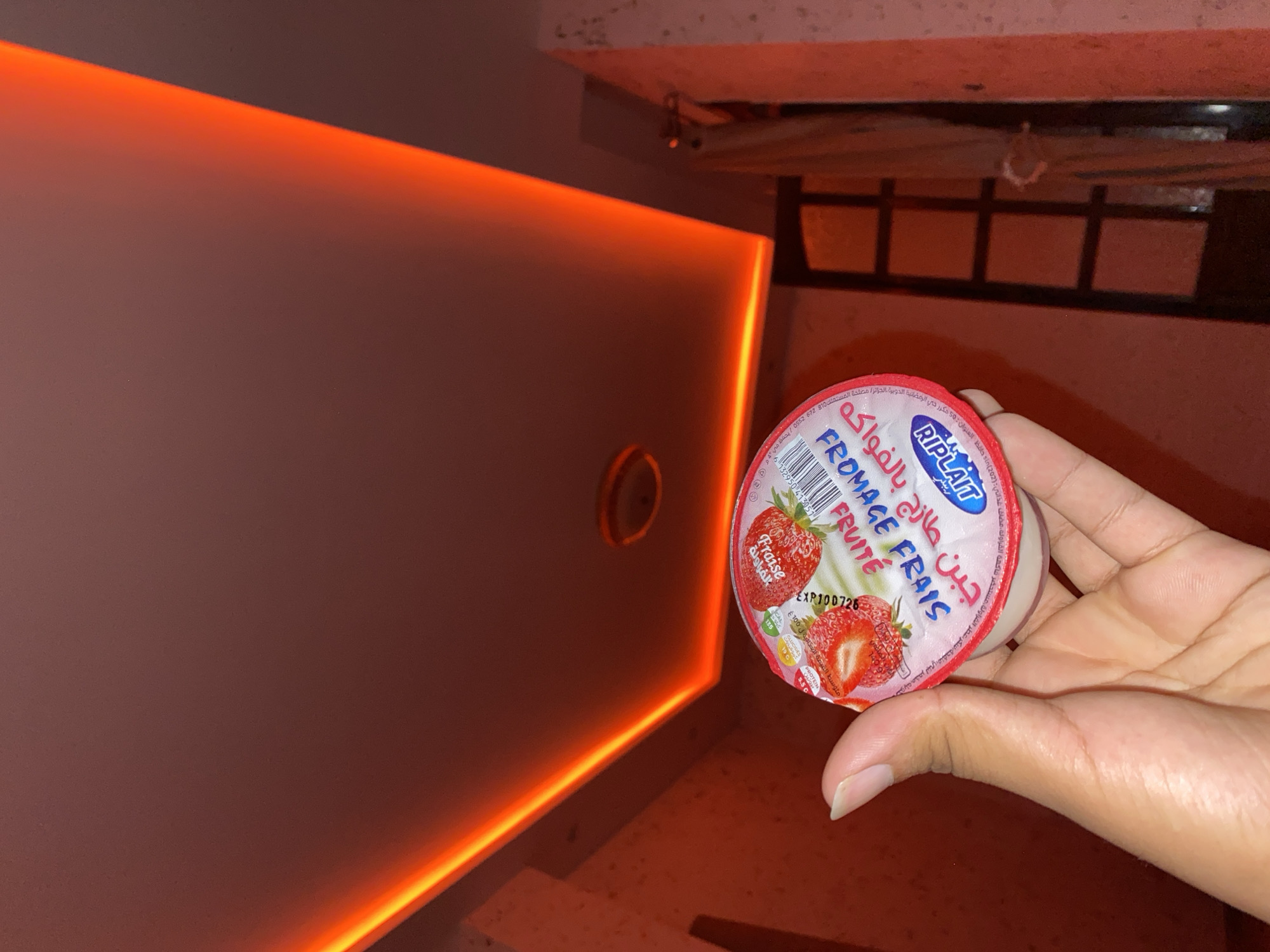 A person's hand, with visible fingernails, holds a yogurt cup with a strawberry design on its lid. The cup is labeled "FROMAGE FRAIS" and "Fruite" in French, and has Arabic text that translates to "strawberry yogurt". The background is a dimly lit room with a ceiling featuring red LED lighting. The overall lighting in the scene is warm and red-toned, likely due to the LED lights. The image is captured from a low angle, looking upwards towards the ceiling. The location is identified as Baraki, Algeria.