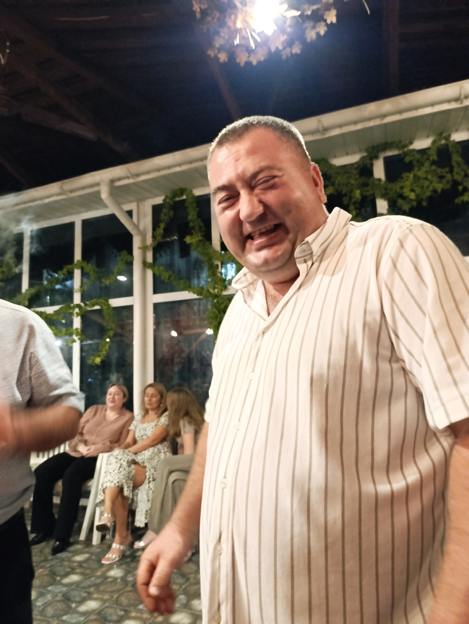 A group of people are gathered outdoors at night. A man in the foreground is smiling broadly, his face close to the camera. He is wearing a light-colored striped shirt. In the background, several women are seated on chairs, also appearing to be enjoying a social gathering. The setting appears to be a veranda or outdoor seating area, with a roof structure overhead and greenery visible through windows. The floor is tiled. The location is Korzhevo, Moldova.