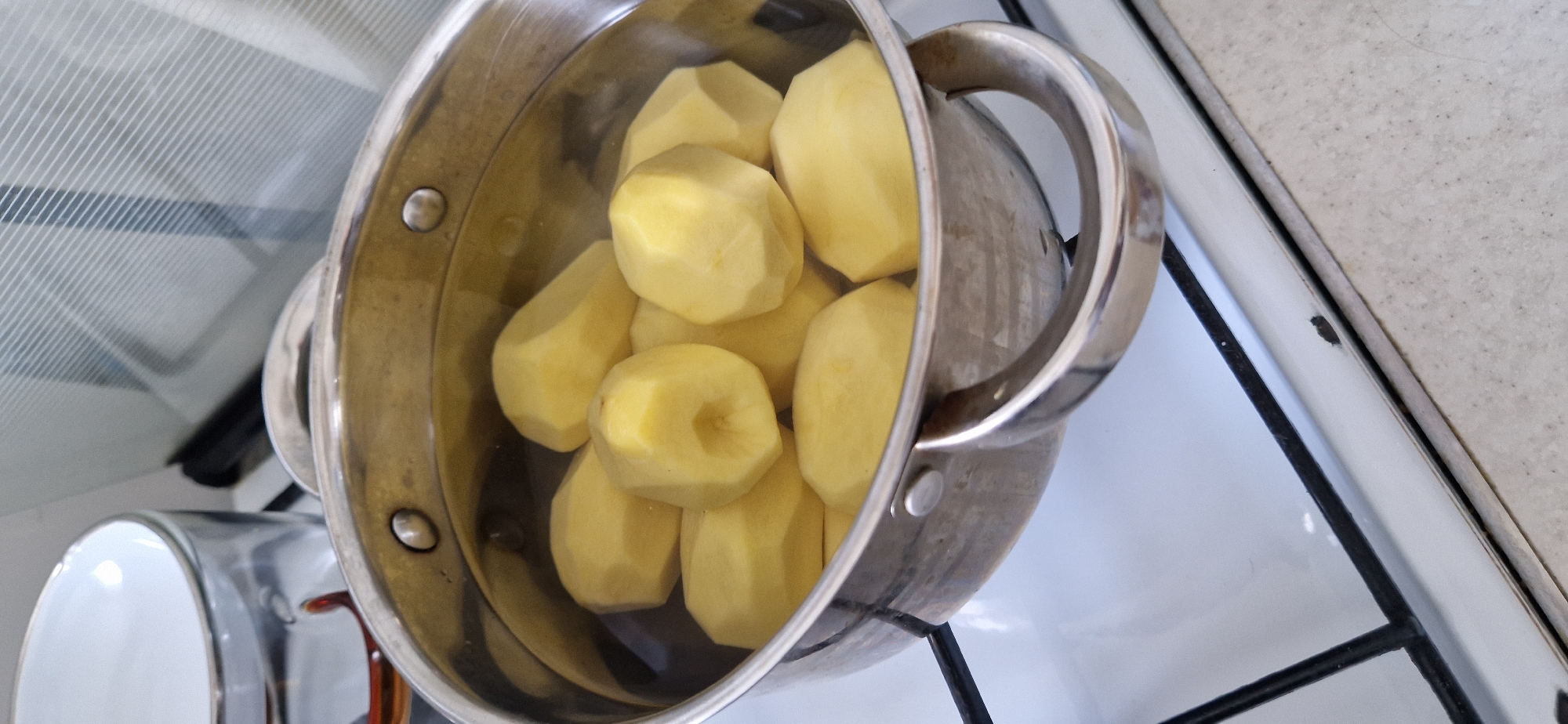 A close-up, overhead shot shows a stainless steel pot filled with peeled potatoes submerged in clear water, sitting on a white gas stovetop. The pot has two riveted handles and is filled with several whole, peeled potatoes. Bubbles are visible on the surface of the water, indicating it is heating. To the left of the pot, a portion of a glass bowl with a white interior and a silver rim is visible. The stovetop is white with black grates. The background is blurred, but appears to be a kitchen environment with a tiled backsplash.