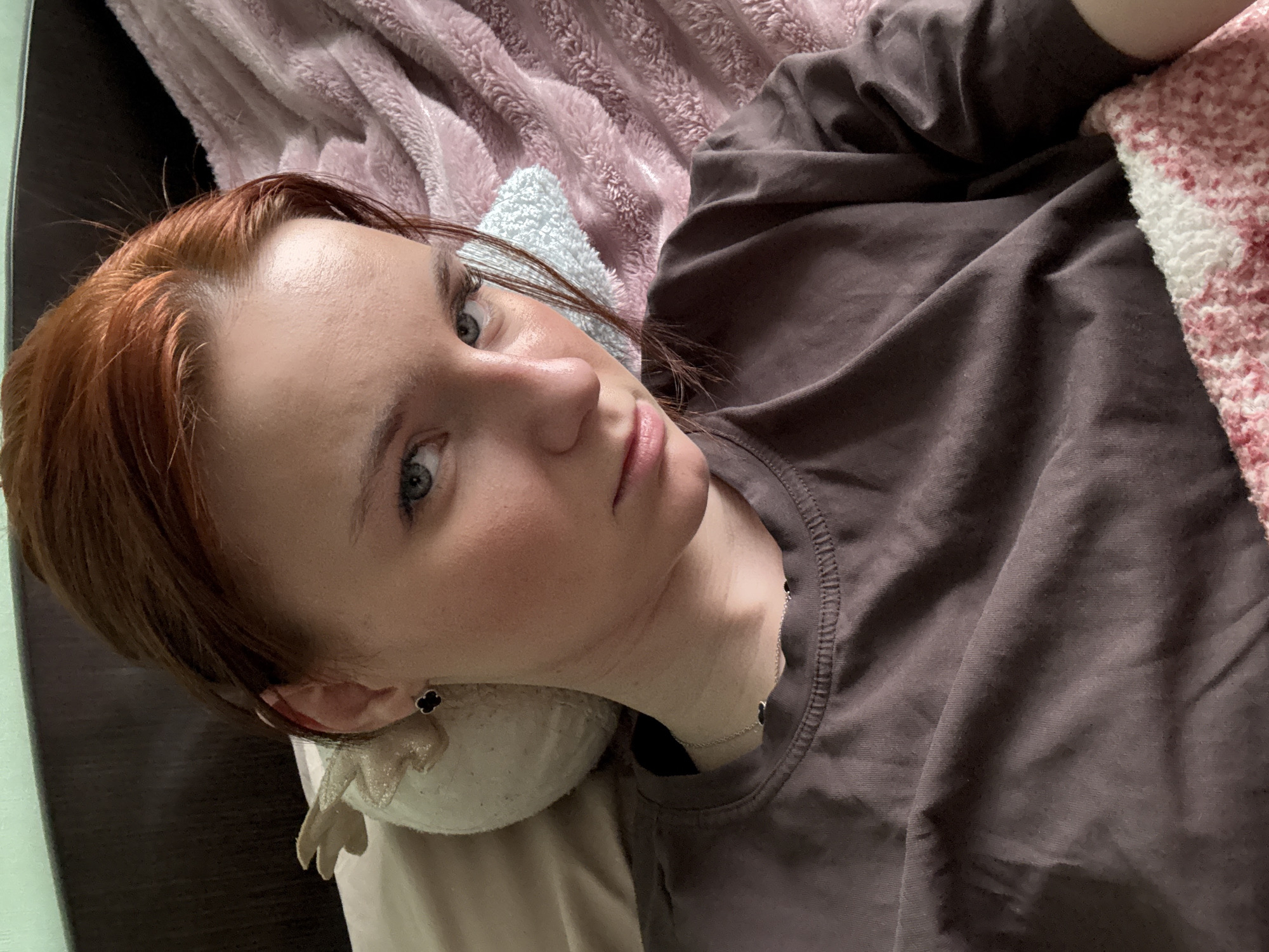A young woman with auburn hair is lying on a bed. She is wearing a brown T-shirt and has light skin. She is resting her head on a soft, off-white pillow. A light pink blanket is partially visible in the background. The scene is indoors, with a dark wooden bed frame visible along the left side of the image. A small black clover earring is visible on her left ear. A dainty silver necklace rests on her chest. The location is unknown, but the city is specified as Unknown, Russia.