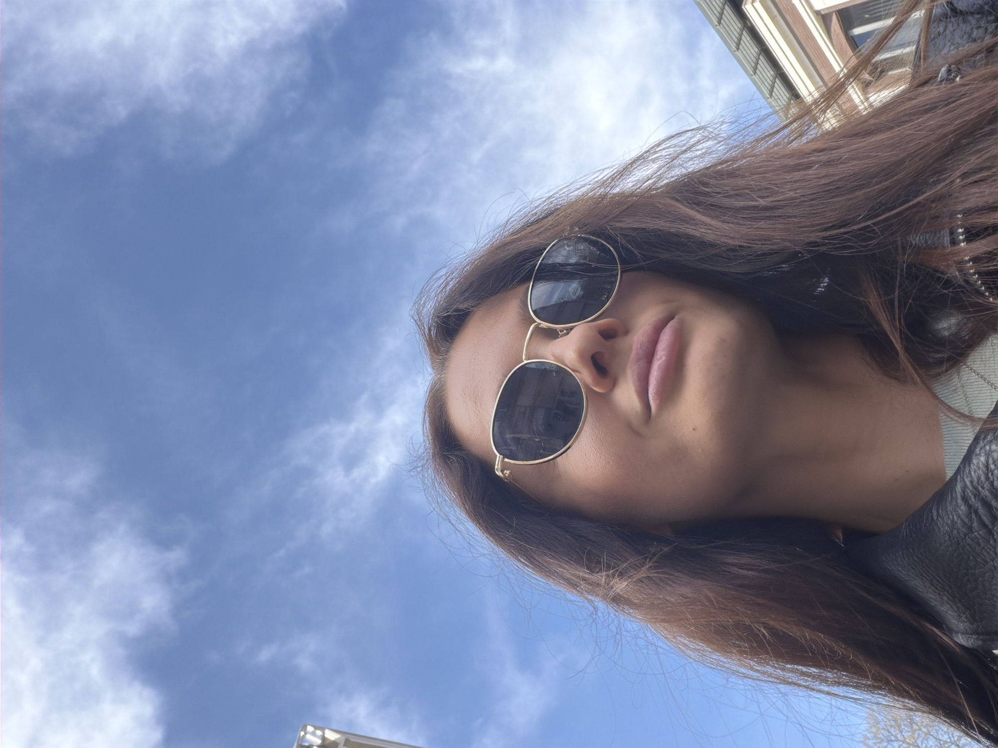 A low-angle, tilted portrait shot captures a person, likely a woman, looking upwards against a bright blue sky with scattered white clouds. The primary subject features long, dark brown hair that cascades down the right side of the frame. She is wearing round sunglasses with gold frames and dark lenses, obscuring her eyes. Her lips are visible, lightly colored in pink and slightly parted. Part of a black, textured garment, appearing to be a leather jacket, is visible on her right shoulder.
The background is dominated by the sky, suggesting an outdoor, daytime setting with clear to partly cloudy weather. In the upper right corner, a partial view of a modern building with white and brown architectural elements is visible. Another subtle architectural detail can be discerned in the bottom left corner, reinforcing an urban environment, consistent with a location like Varna, Bulgaria. The image is composed at a dynamic angle, creating an impression of the subject gazing towards the sky or upward at the camera. There is no visible text in the image.