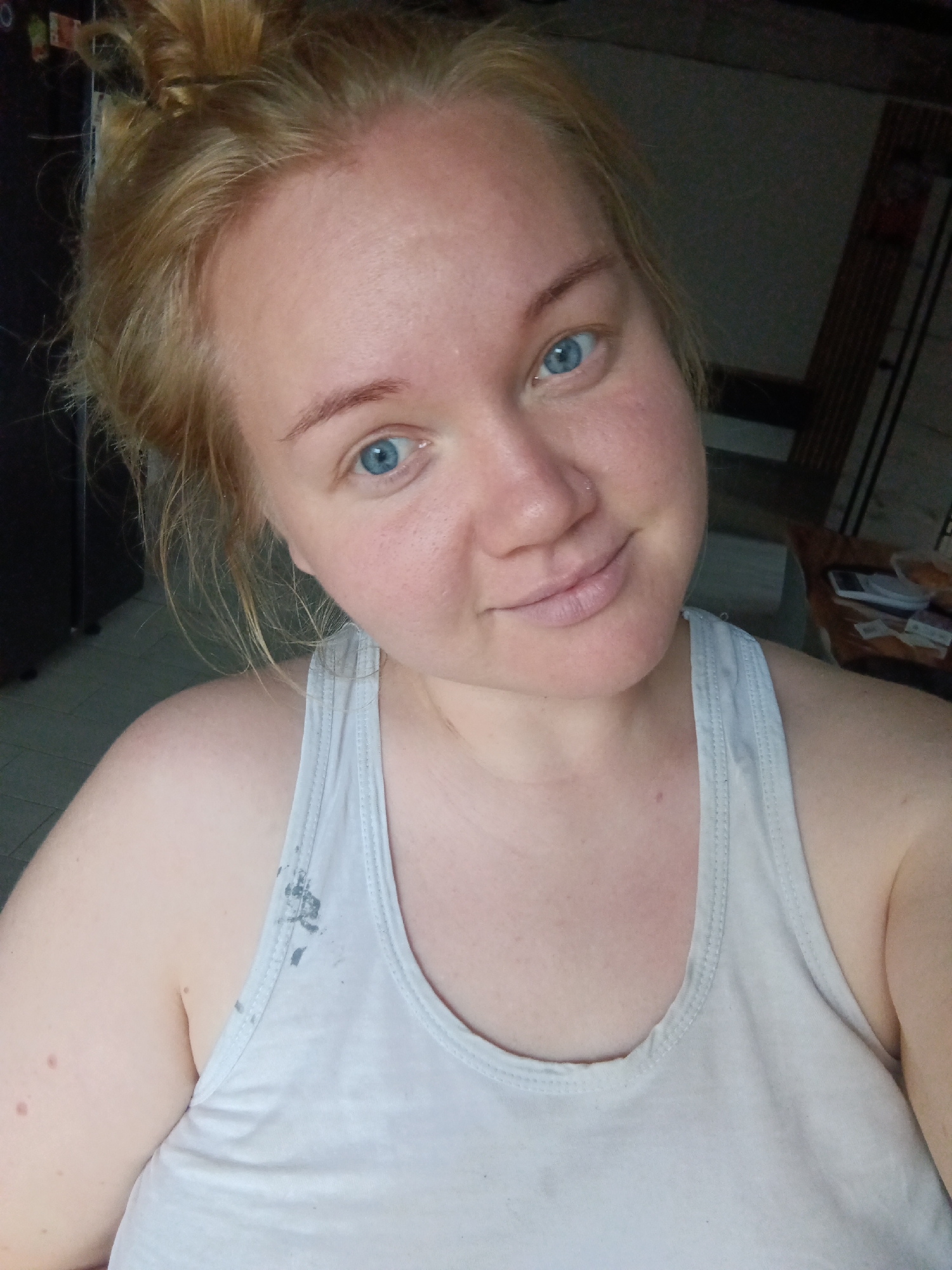 A medium shot shows a fair-skinned woman with blonde hair in a messy bun, blue eyes, and a nose ring, looking directly at the camera and smiling slightly. She is wearing a white tank top with some dark smudges on it. The background is dimly lit, with a dark wall on the left, some shelving units on the right, and a tiled floor. The overall impression is of a casual, indoor setting, possibly a home. The image quality is slightly grainy. There is no visual information to place the image in Фоки, Russia.
