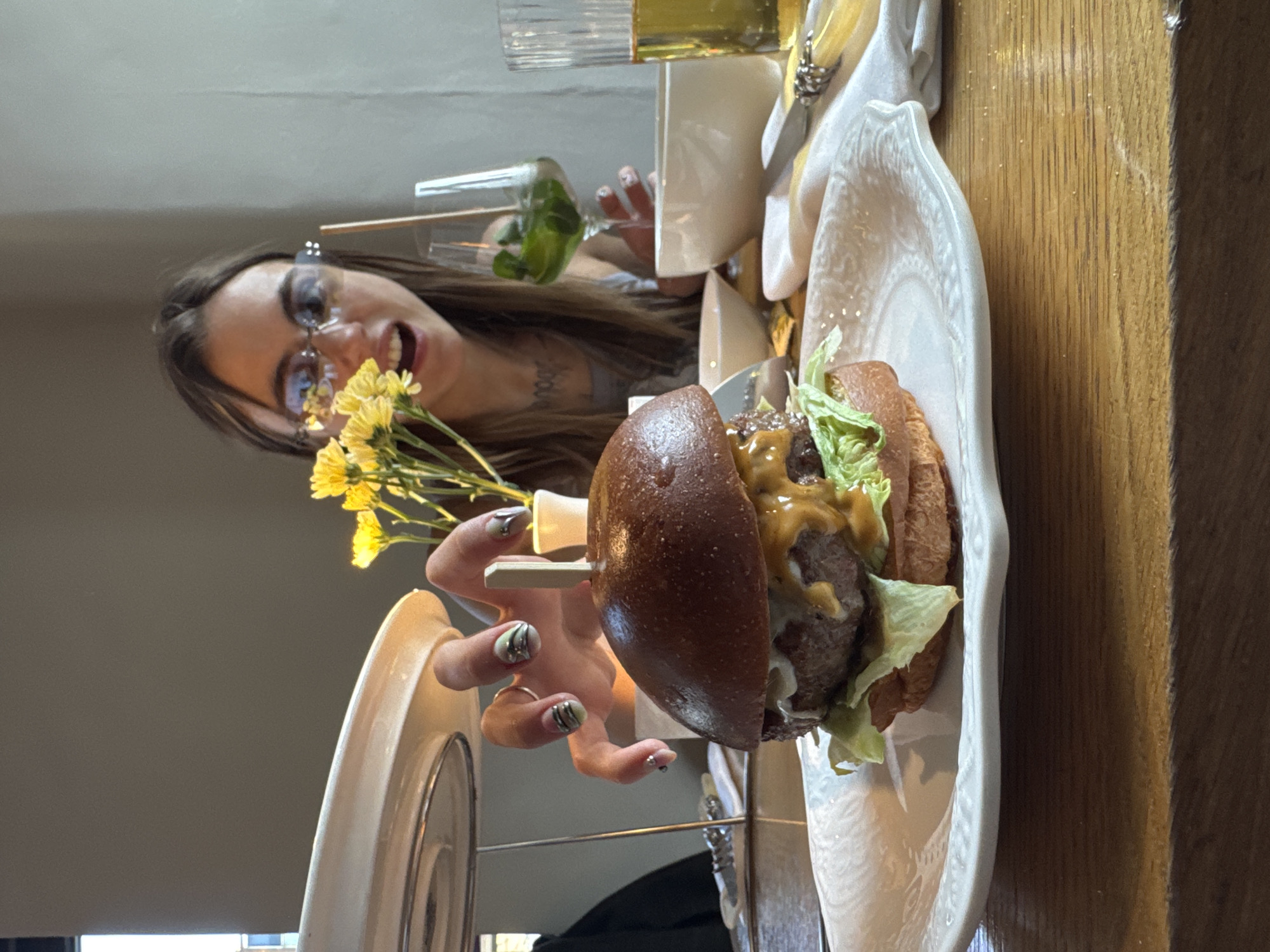 The image, captured in Moscow, Russia, depicts a lively dining scene centered around a young woman and a gourmet burger. A young woman with long, dark hair and modern glasses is positioned in the foreground, gazing towards the viewer with an open mouth, conveying an expression of surprise, excitement, or amusement. Her left hand is visible, holding a small bunch of yellow flowers, and showcasing meticulously painted fingernails with a greenish-silver base and black wavy designs.
In the mid-ground, a beautifully presented gourmet burger sits on a distinctive white, wavy plate. The burger features a dark bun, a substantial meat patty, crisp green lettuce, and a rich yellowish sauce, held vertically by a white skewer. Various drinks are visible in the background: a tall glass containing a clear liquid and another glass with a green garnish (likely mint) and a straw, suggesting a cocktail. Elegant white tableware, including a napkin adorned with a decorative silver ring, completes the setting. The scene unfolds on a warm-toned wooden table, under what appears to be daytime ambient lighting, within a restaurant or cafe environment. The overall impression is that of a cheerful and engaging moment during a meal. No visible text is present in the image.