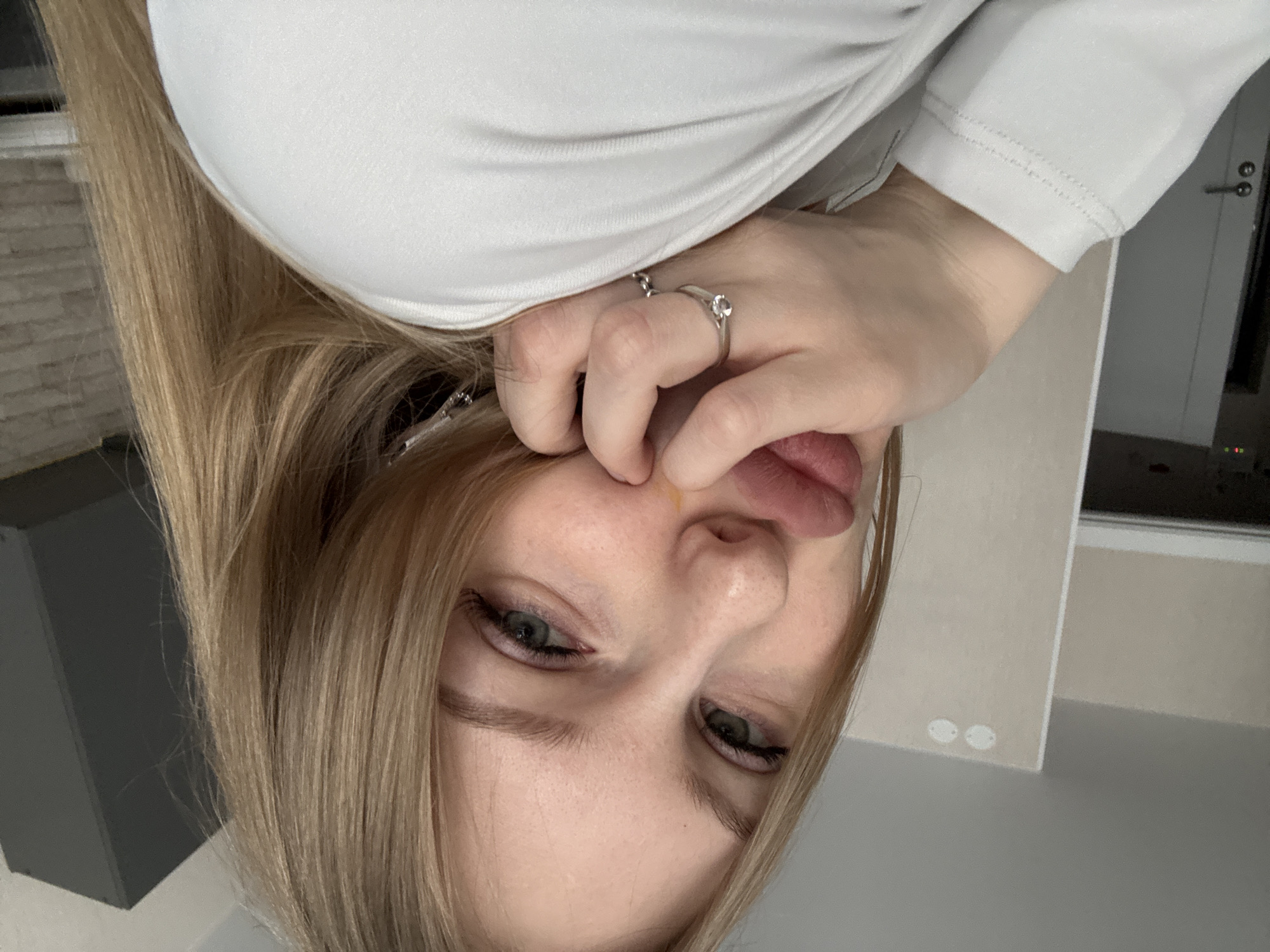 A close-up, upside-down selfie of a Caucasian woman with blonde hair and green eyes. She is wearing a light-colored, long-sleeved shirt and two rings on her fingers. She is holding the bridge of her nose with her fingers. Her tongue is slightly out. The background shows a blurred interior with a door and a wall. The location in Perm, Russia is not evident in the image.