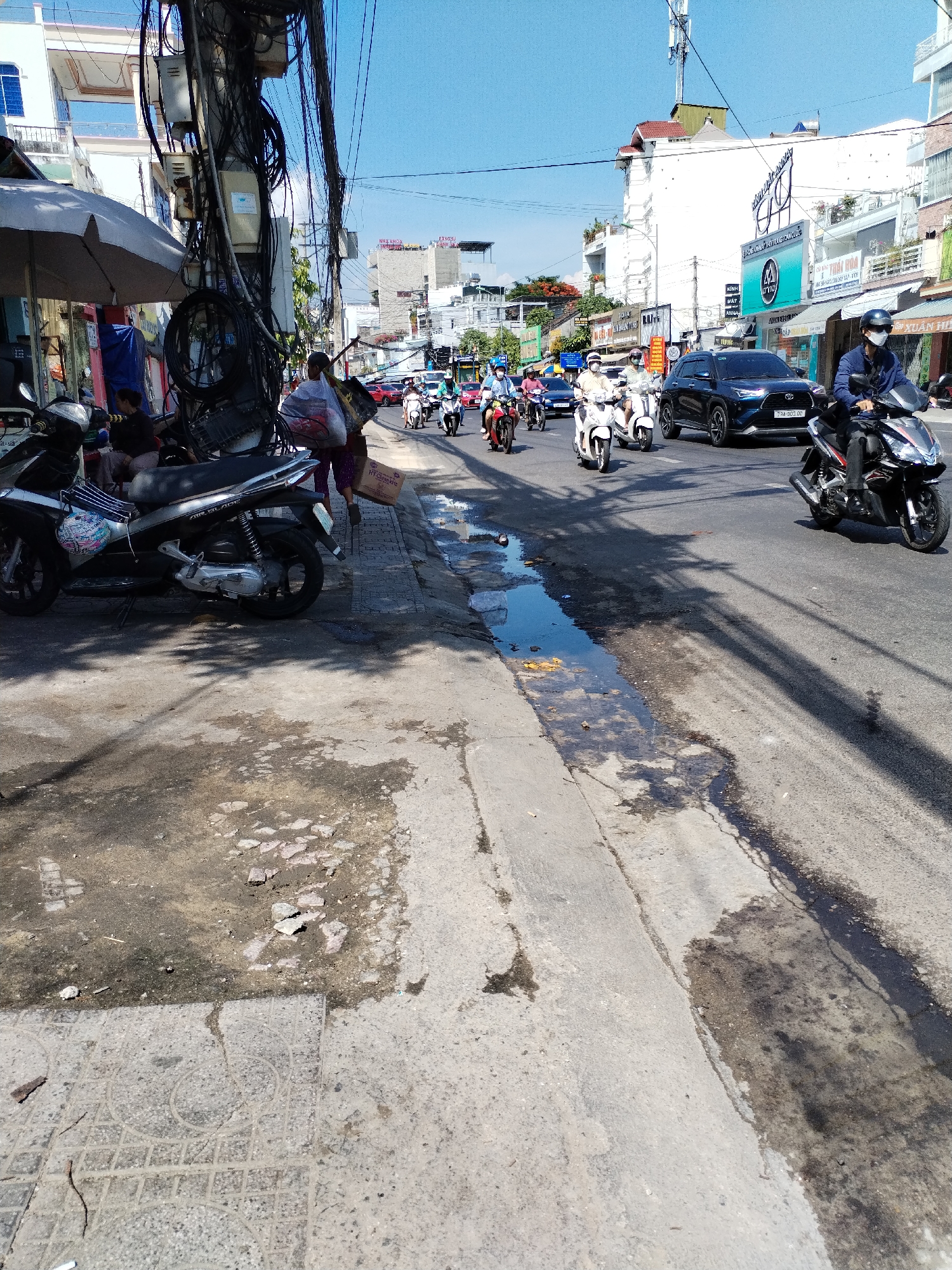 The image depicts a street scene in Bac Nha Trang Ward, Vietnam. A busy road is filled with motorcycles and a few cars. On the left side of the road, there is a sidewalk with a parked motorcycle and some people standing. A large cluster of electrical wires hangs overhead. The road surface appears worn and there are puddles of water near the curb. Buildings line the opposite side of the street, with signs indicating commercial establishments. The sky is clear and blue, suggesting a sunny day.