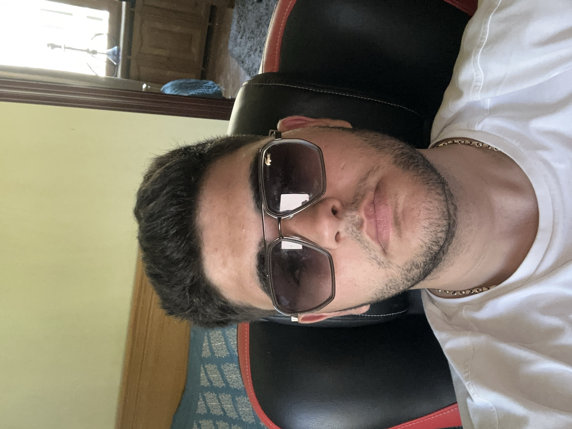 A man wearing sunglasses and a white t-shirt sits in a black and red chair. He has short dark hair, a beard, and a gold chain around his neck. The location is indoors. The city and country are not discernible from this image.