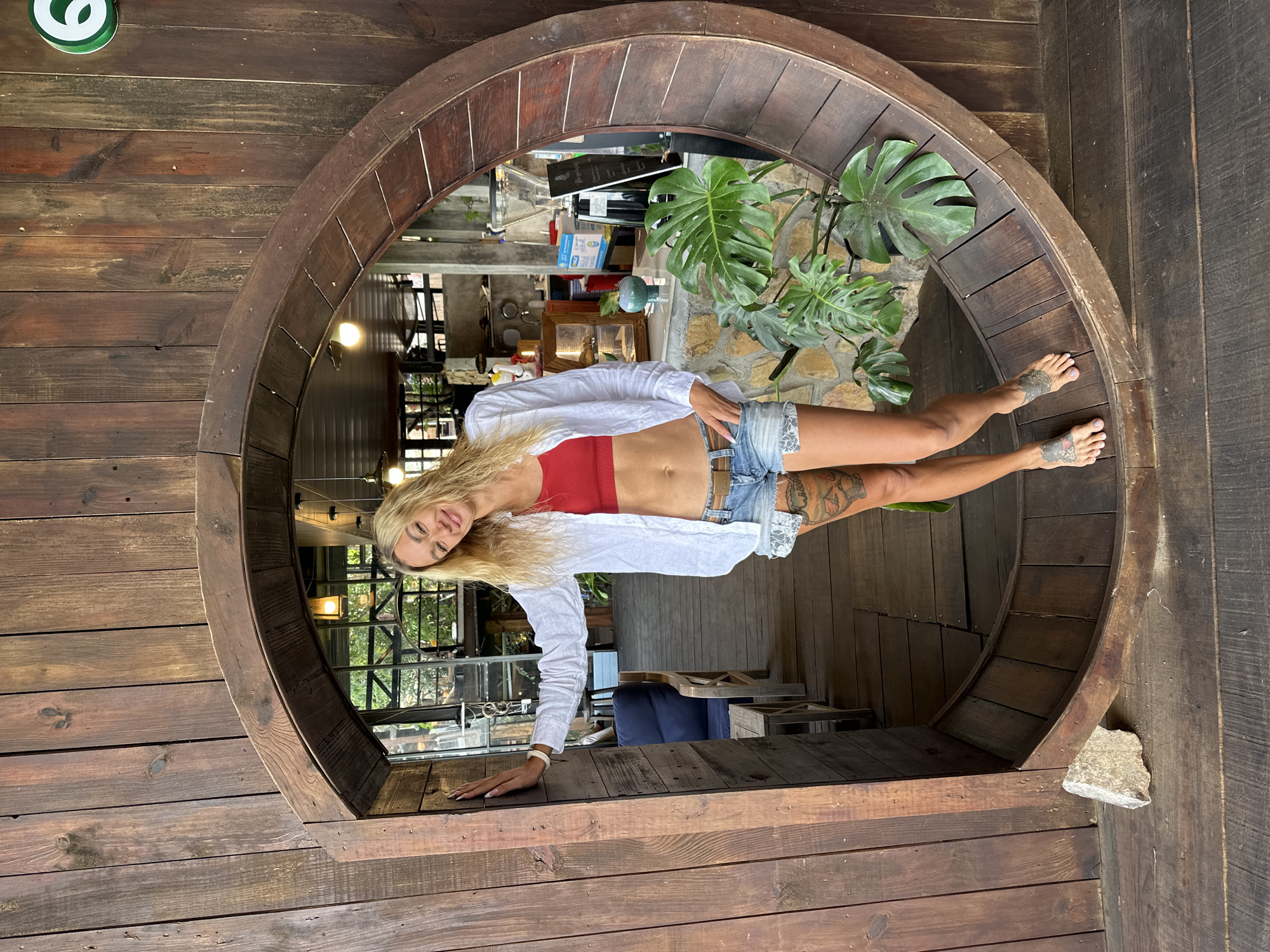 A woman with blonde hair and tattoos on her legs is standing in a circular wooden archway. She is wearing a red crop top, a white open shirt, and denim shorts. The archway is set against a wooden wall. Behind the woman, a cafe is visible with tables, chairs, and a counter. Plants are also present in the scene. The woman is smiling and looking towards the camera. The location is stated to be in Da Lat, Vietnam.