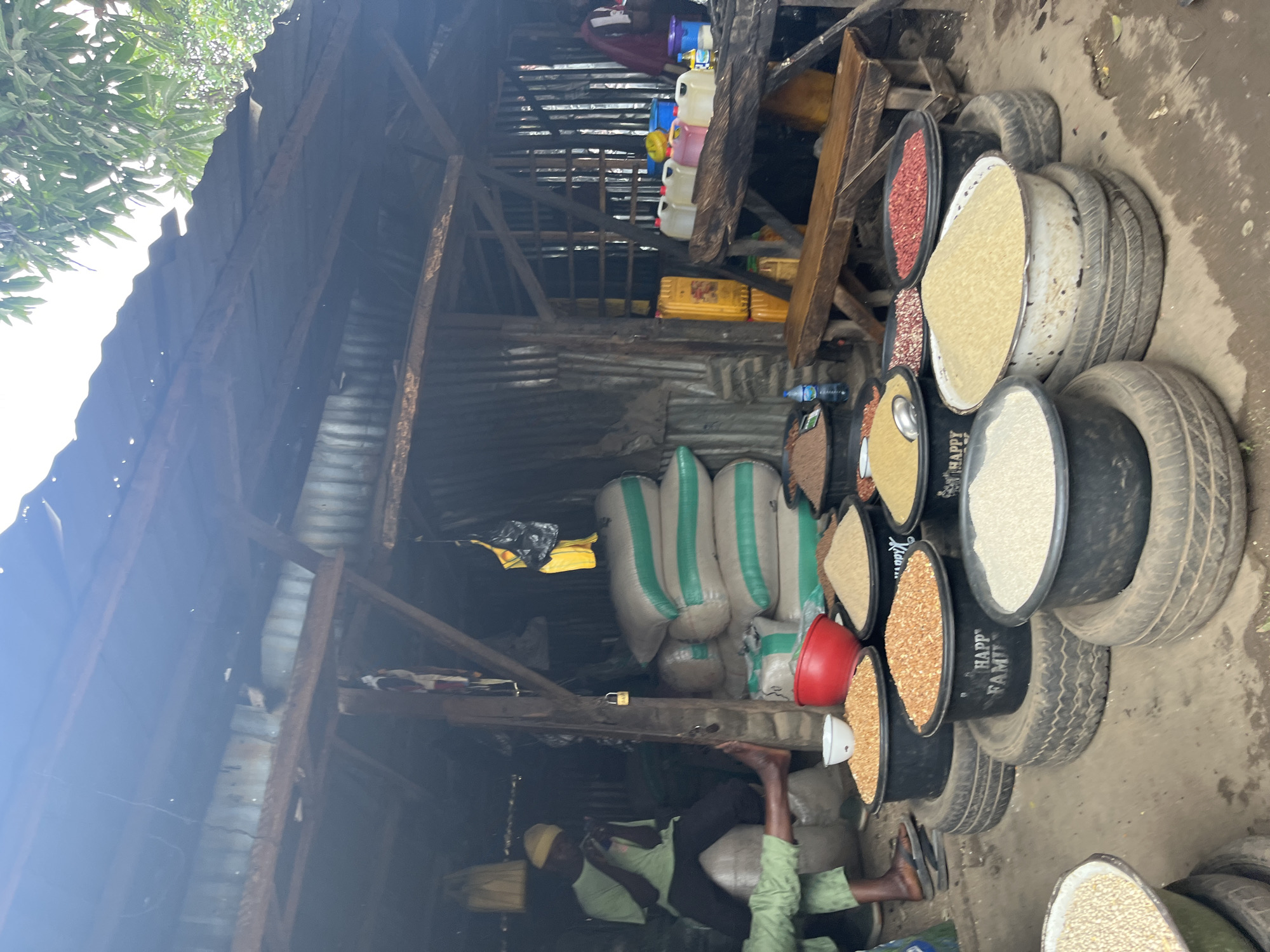 This image depicts an outdoor market stall in Jos, Nigeria, likely selling grains and food items. In the background, a person wearing a green t-shirt and yellow cap is seated, seemingly engaged with a mobile phone. Several large sacks, possibly containing grains, are stacked prominently, marked with horizontal green stripes. The stall's structure is rustic, featuring a corrugated metal roof supported by wooden beams, and walls made of corrugated metal sheeting.
In the foreground, various large containers, some resting on stacked tires, are filled with different types of grains. These include what appears to be corn, rice, and possibly beans or lentils, distinguished by their varying colors and textures. Red and white bowls are also visible among the containers. To the right, a wooden bench is positioned in front of a table laden with plastic containers of different sizes and colors, some containing liquids and others appearing to be foodstuffs. Yellow jerrycans and water bottles are also present. The ground is dirt and appears wet in places. The overall scene suggests a bustling local market environment.