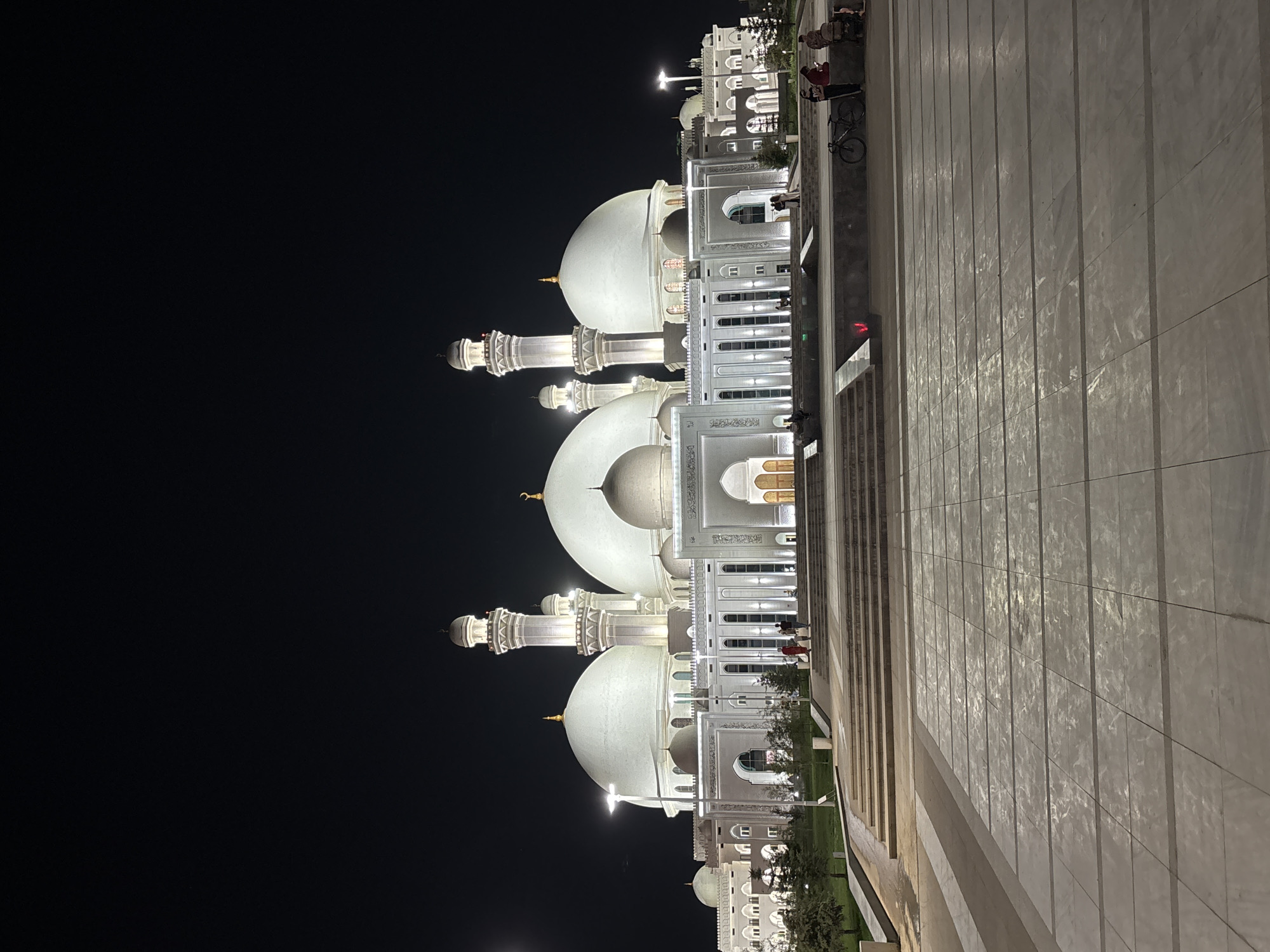 An exterior, low-angle, full shot at night captures the grandeur of the Hazrat Sultan Mosque in Shymkent, Kazakhstan. The mosque, illuminated by artificial lights, dominates the frame with its white domes and minarets set against a black sky. The architectural style is Islamic, characterized by multiple large domes and slender, tall minarets. The building is expansive, with a symmetrical facade and numerous windows. In the foreground, a large, tiled plaza stretches out, leading up to a set of wide stone steps. Several people are visible on the steps and the plaza, some appearing to be conversing or resting, while others are walking. A bicycle is parked near a group of people on the plaza. The overall atmosphere is one of quiet reverence and impressive scale.