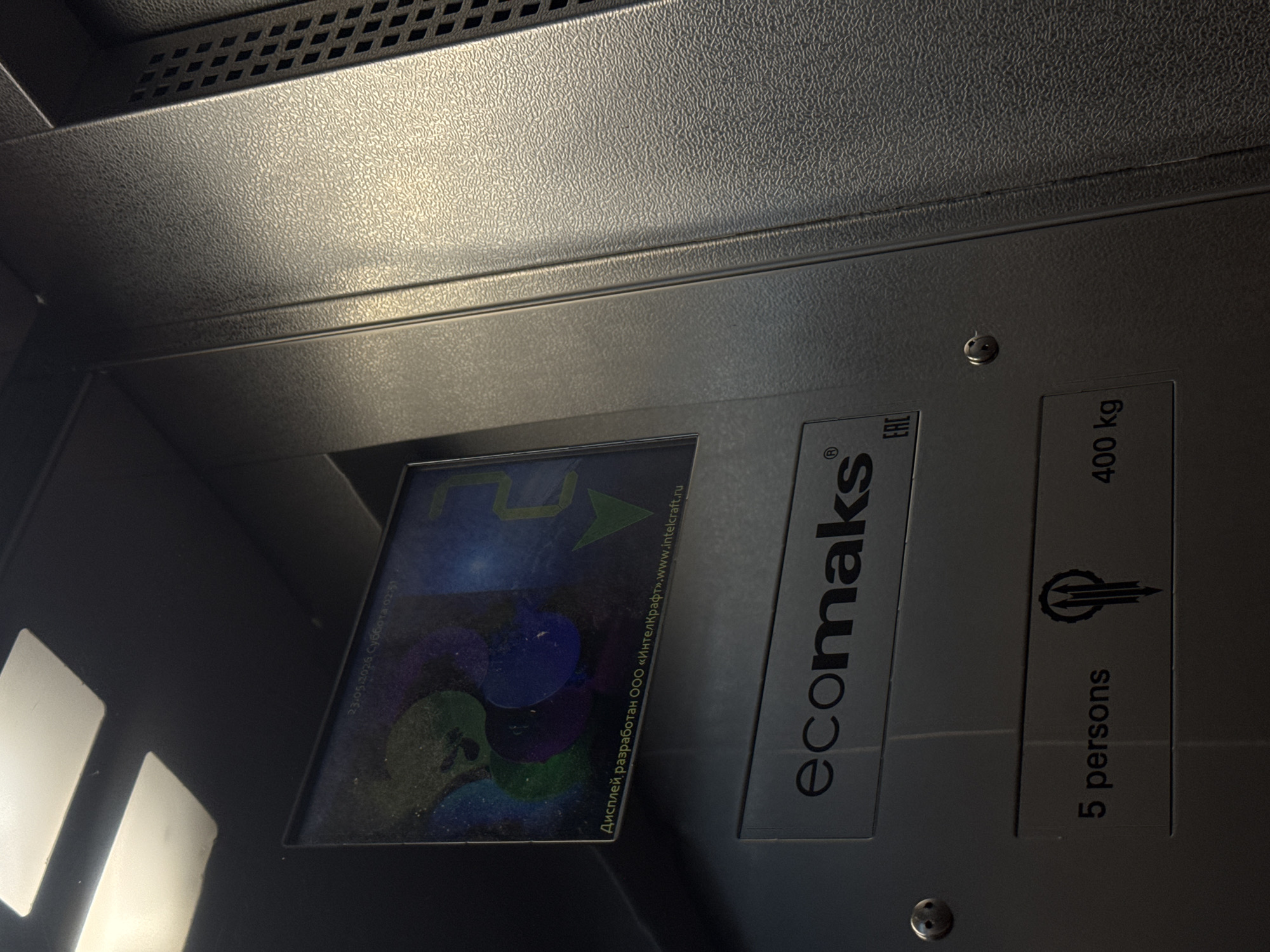 The image shows a close-up of an elevator panel. The panel has a digital display showing a graphical interface and some text. To the right of the display, there is labeling including "ecomaks" in lowercase, along with the symbols EAC and a logo. Further to the right, the label indicates that the elevator can hold "5 persons" and has a maximum weight capacity of "400 kg". The text on the digital display is in Russian and includes a date, "23.05.2026", and the statement that the display was developed by "IntelKraft". The panel is gray. The location, Yevpatoriya, Ukraine, is not discernible from the image alone.