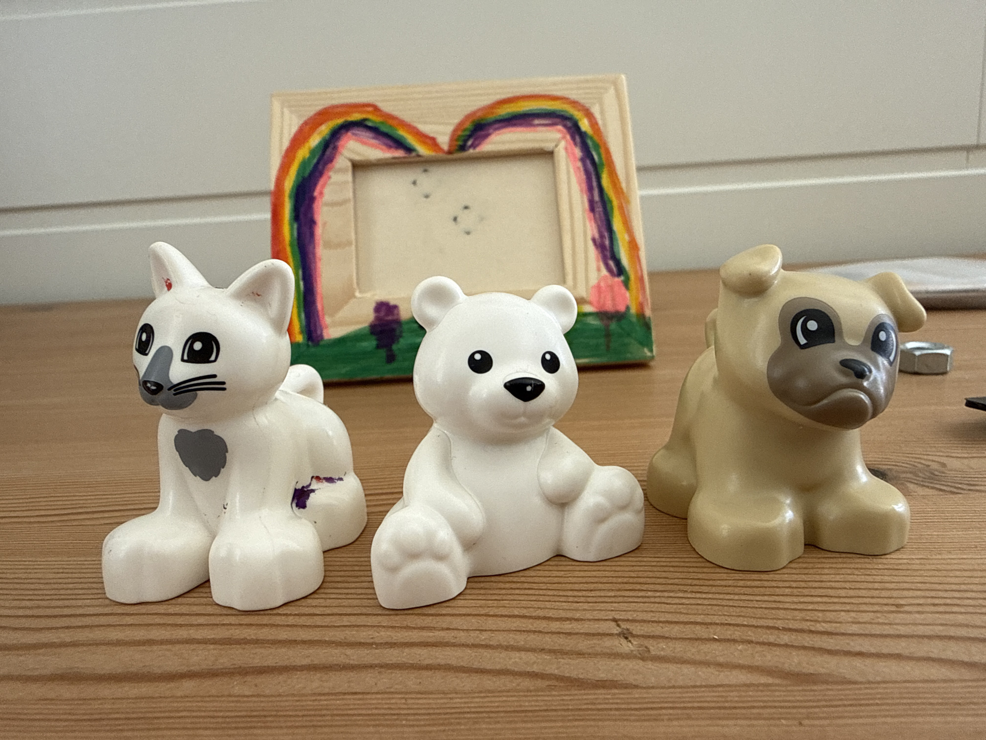 Three plastic animal figurines, a white cat, a white polar bear, and a tan pug, are positioned on a wooden surface. Behind them, a wooden picture frame is decorated with a colorful rainbow and green landscape. The cat has a grey heart on its chest and a smudge of purple on its side. The pug has a dark brown muzzle. The background shows a white wall and possibly a door. The scene is set indoors, on a wooden table, in what appears to be a home environment. There are no people visible in the image. The location in the city of Ridabu, Norway is not discernible from the image.