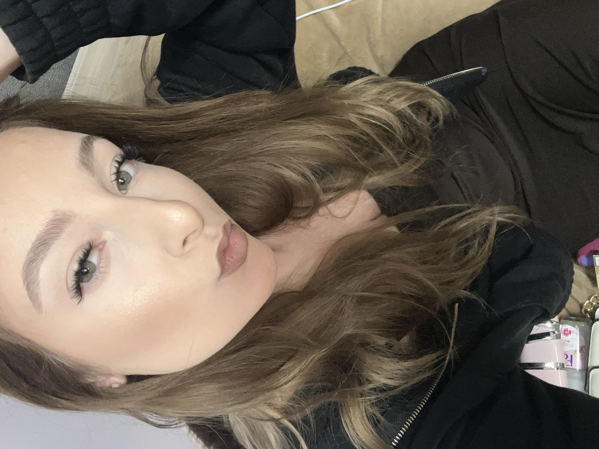 A close-up, tilted selfie shows a young woman with light skin, light brown hair, and green eyes. She is wearing makeup including false eyelashes, eyebrow shaping, and lip color. She's wearing a black zip-up hooded sweatshirt. The background includes a beige surface. The image was not taken in Varna, Bulgaria.