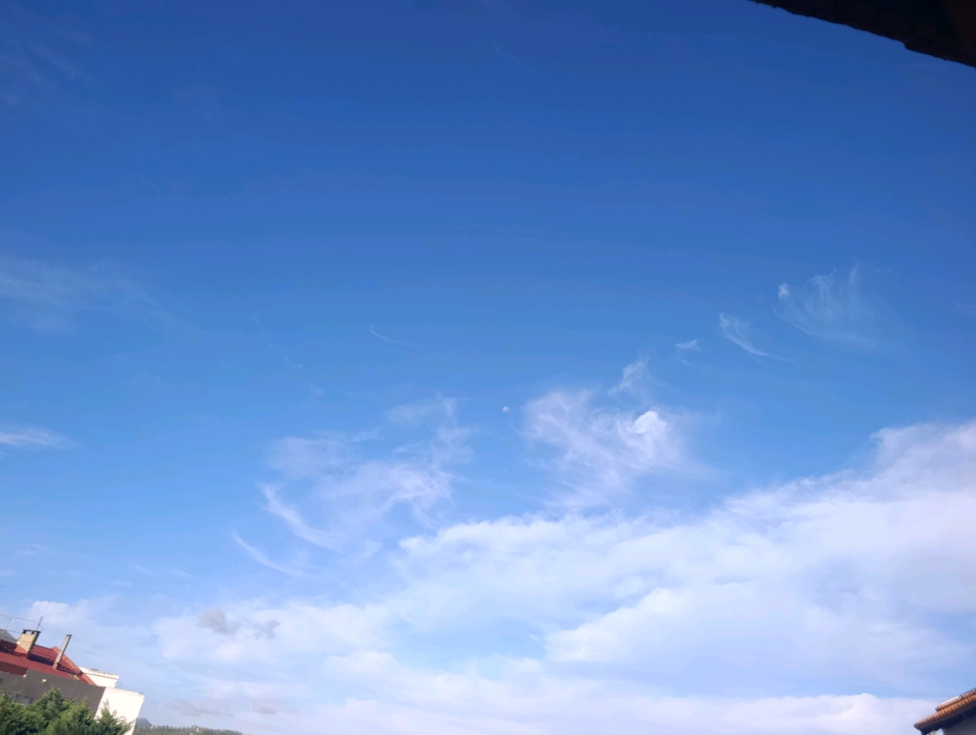 The image captures a bright, clear blue sky with wispy cirrus clouds scattered across it. The lower portion of the image reveals the rooftops of buildings in Setúbal, Portugal, with red-tiled roofs and some white structures visible. Green foliage can be seen in the bottom left corner, suggesting a residential or urban area with some vegetation. A dark, possibly wooden or metal overhang, is present at the top edge of the frame, suggesting a view from a balcony or window. The overall scene is serene and depicts a typical daytime view in the city.